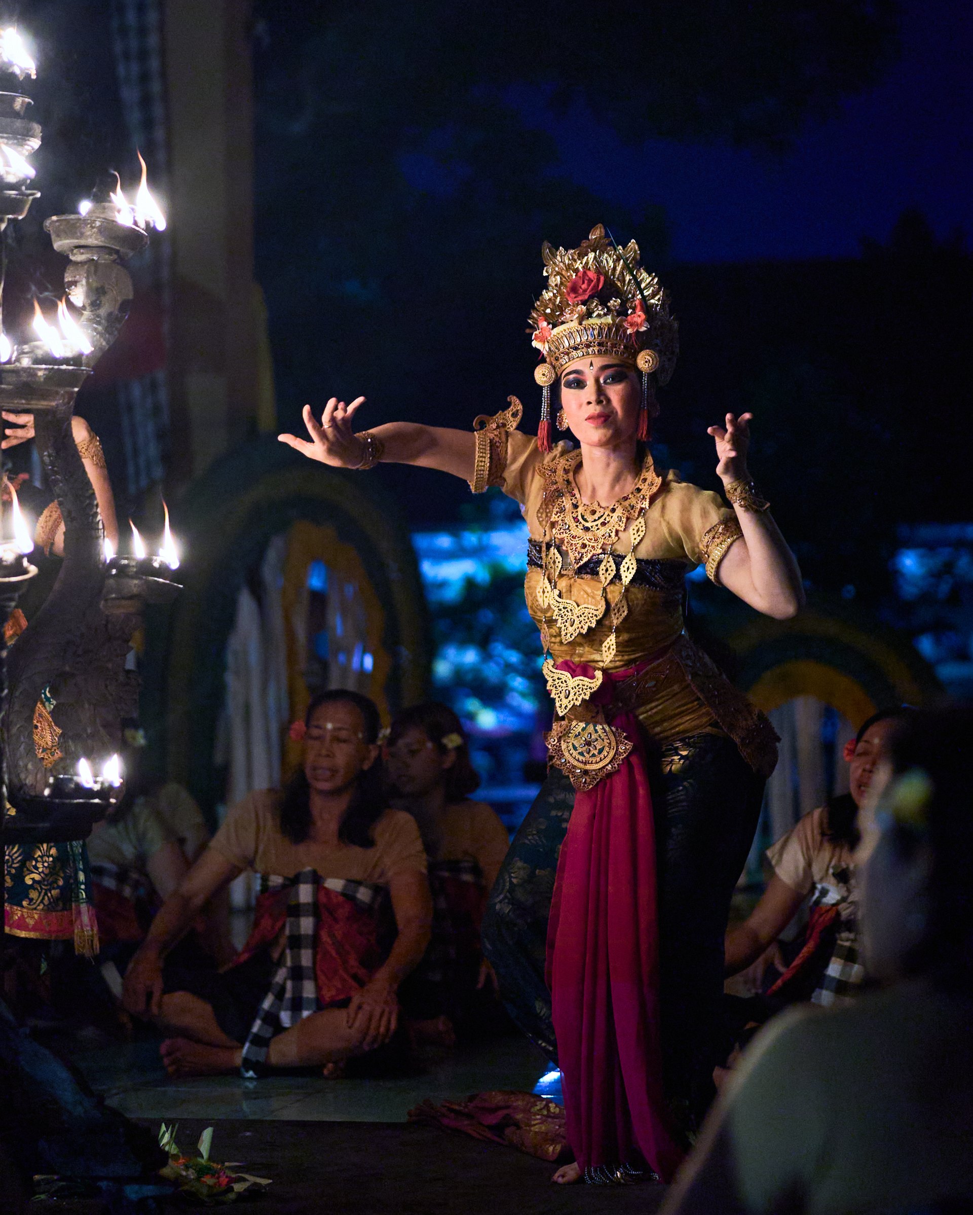 Balinese dancer in traditional ceremonial dress with gold ornaments and headdress performing at night with theatrical stage lighting and an audience visible below.