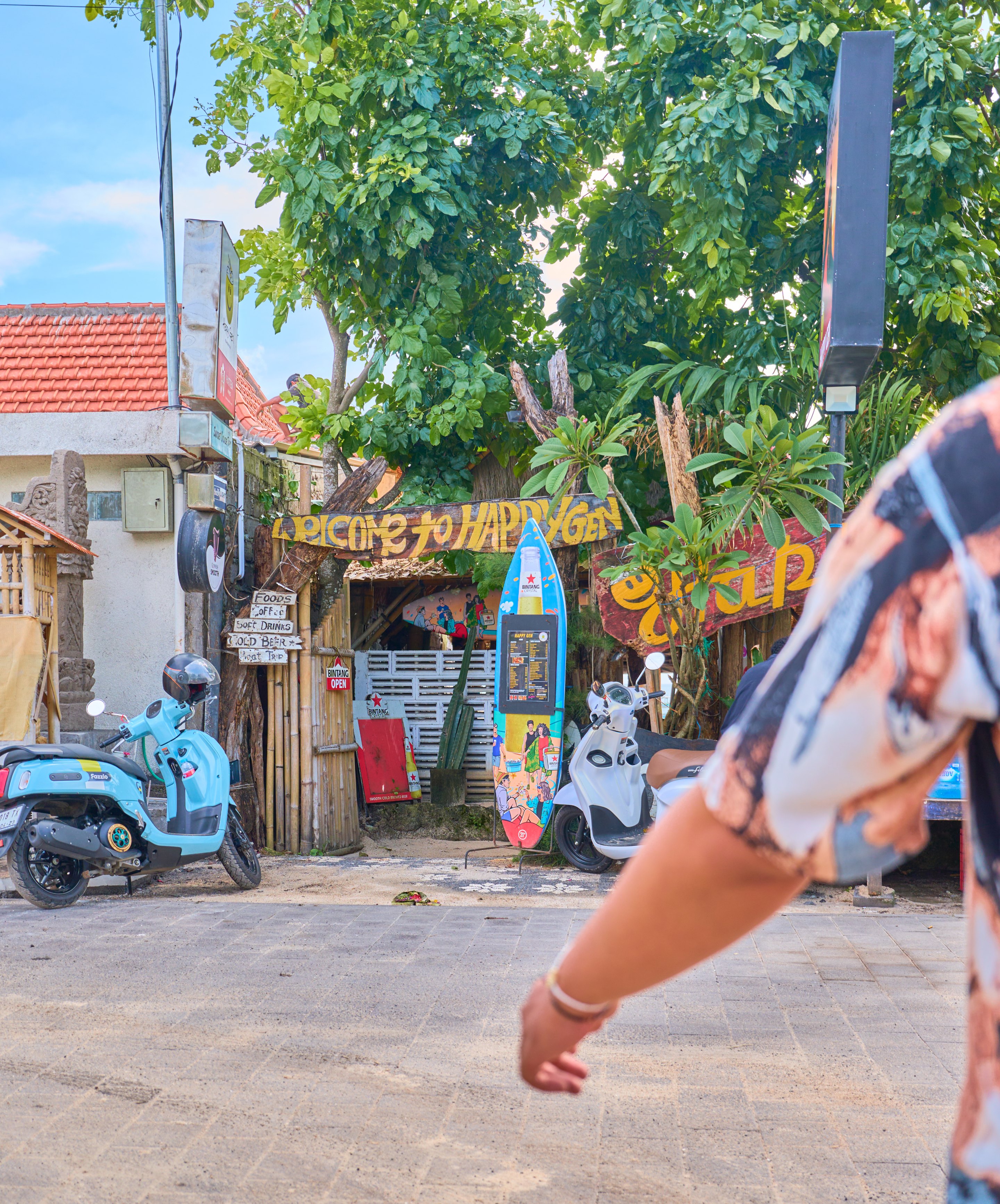 Street scene with a blue scooter parked on the left, colorful storefront signage and decorations, and a person's arm in motion in the foreground.