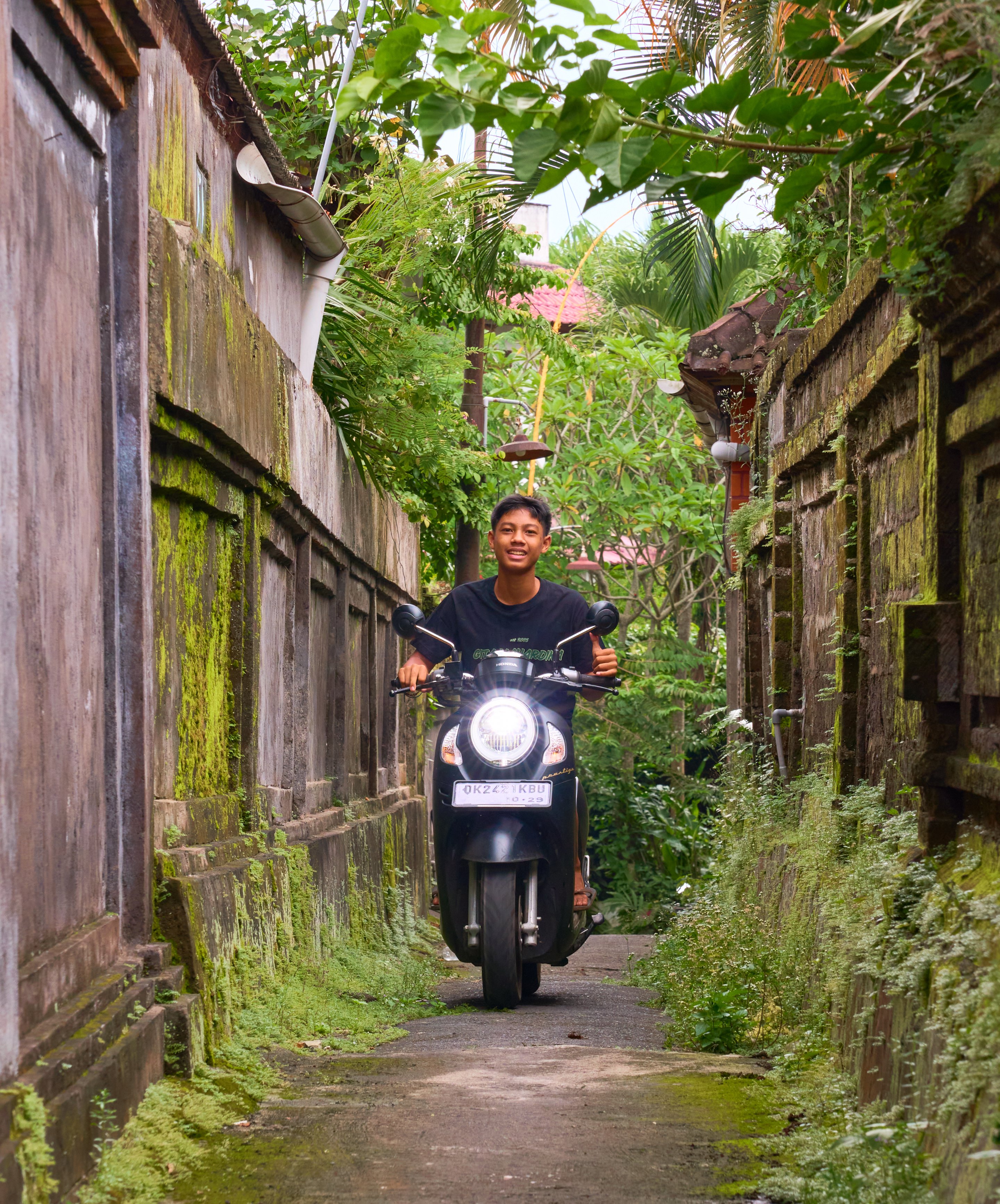 Man riding a motorcycle down a narrow moss-covered alley with weathered stone walls on both sides, dense tropical foliage overhead, and the scooter's headlight visible.