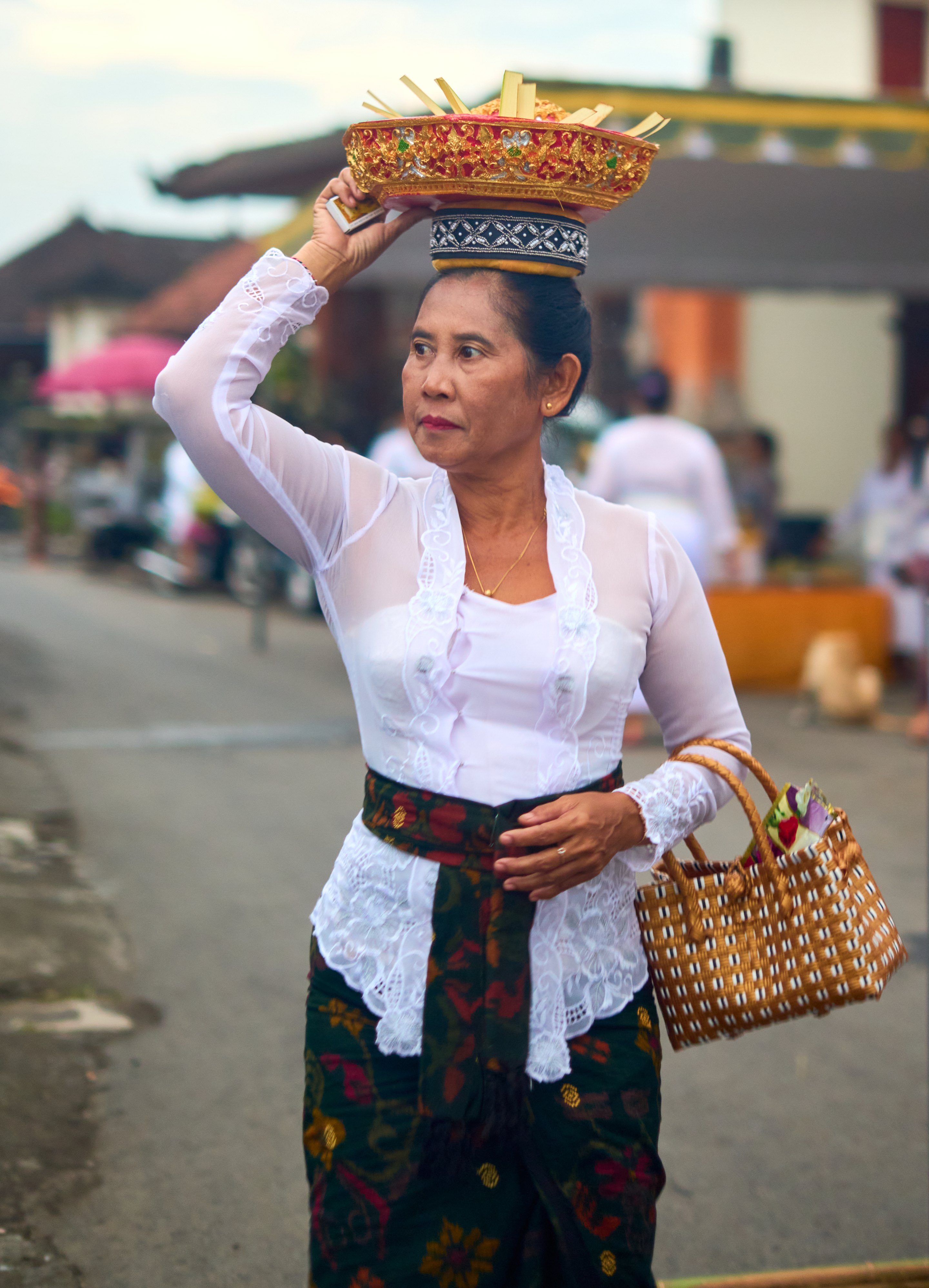Woman wearing traditional Balinese dress with a white long-sleeved top, decorative waist belt, patterned sarong, ornate crown, and woven basket, standing on a street.