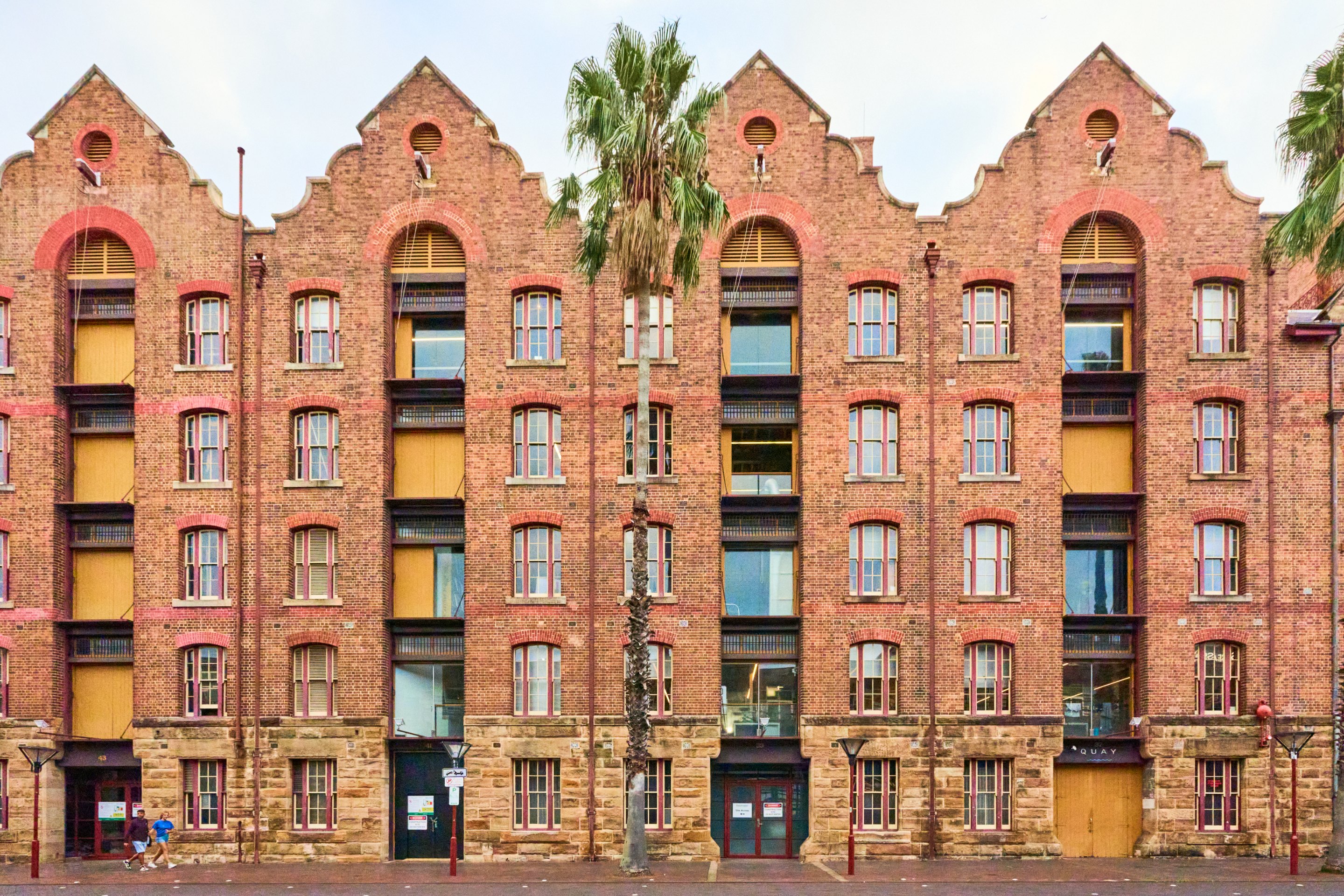 Historic red-brick warehouse building with distinctive Dutch-style stepped gable roofline, large arched windows and doorways, palm tree on right.