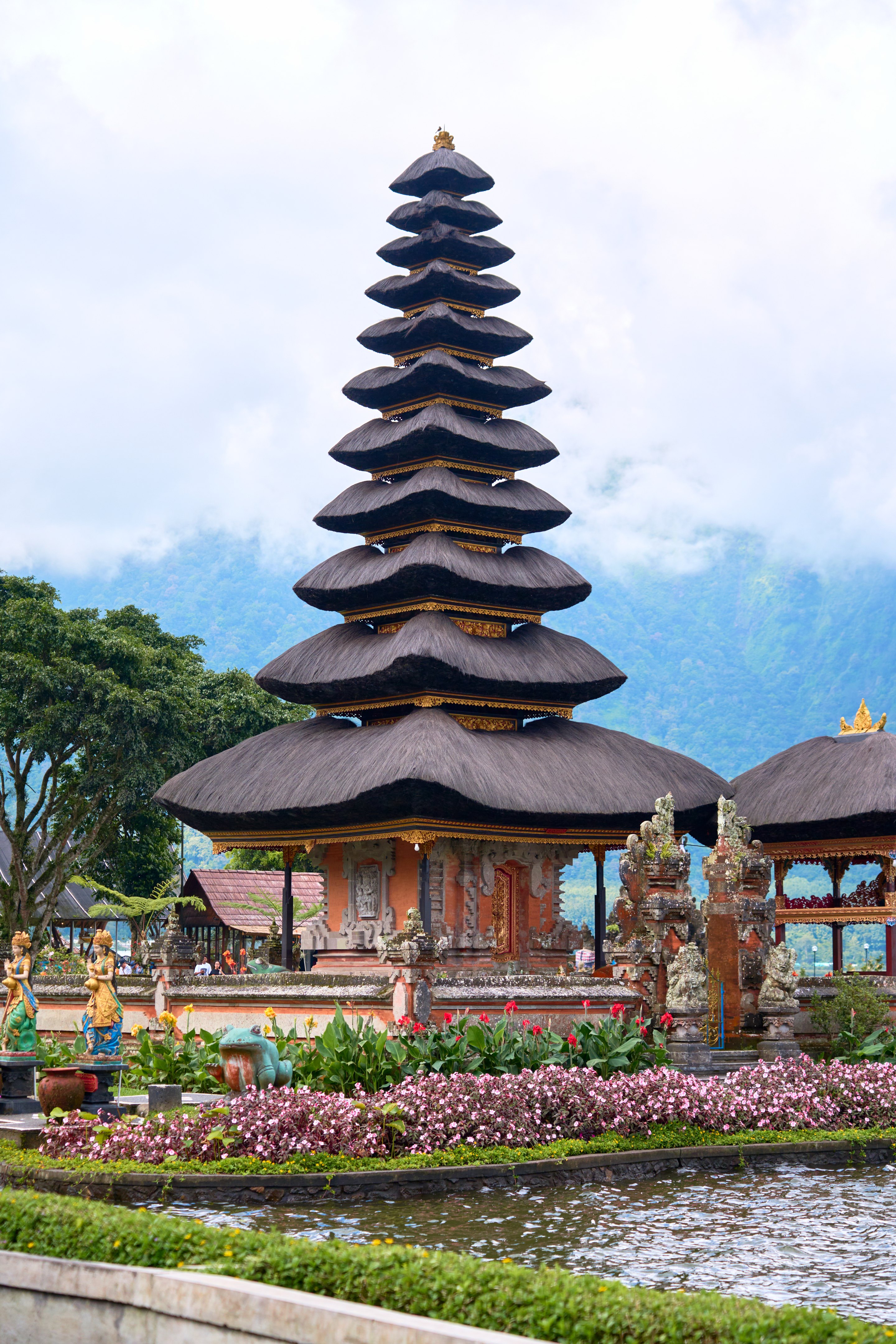 Balinese temple with distinctive tiered pagoda-style dark roofs reflected in a pool, surrounded by purple blooming flowers and trees, with mountains visible behind.