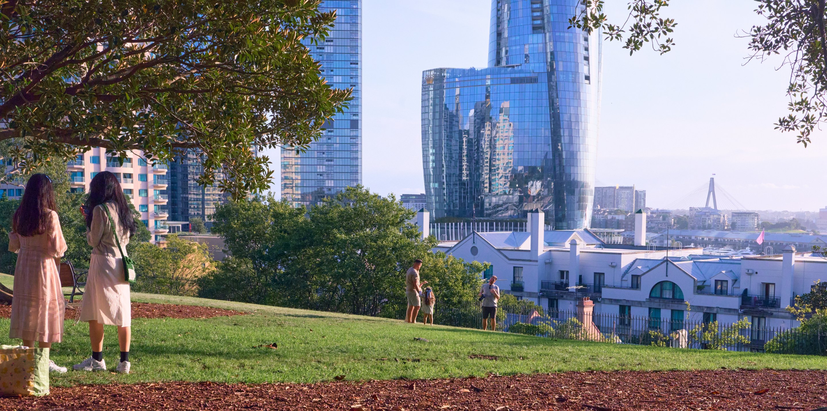 Two people standing on grassy hillside park overlooking city skyline with tall buildings, trees framing edges, clear blue sky.
