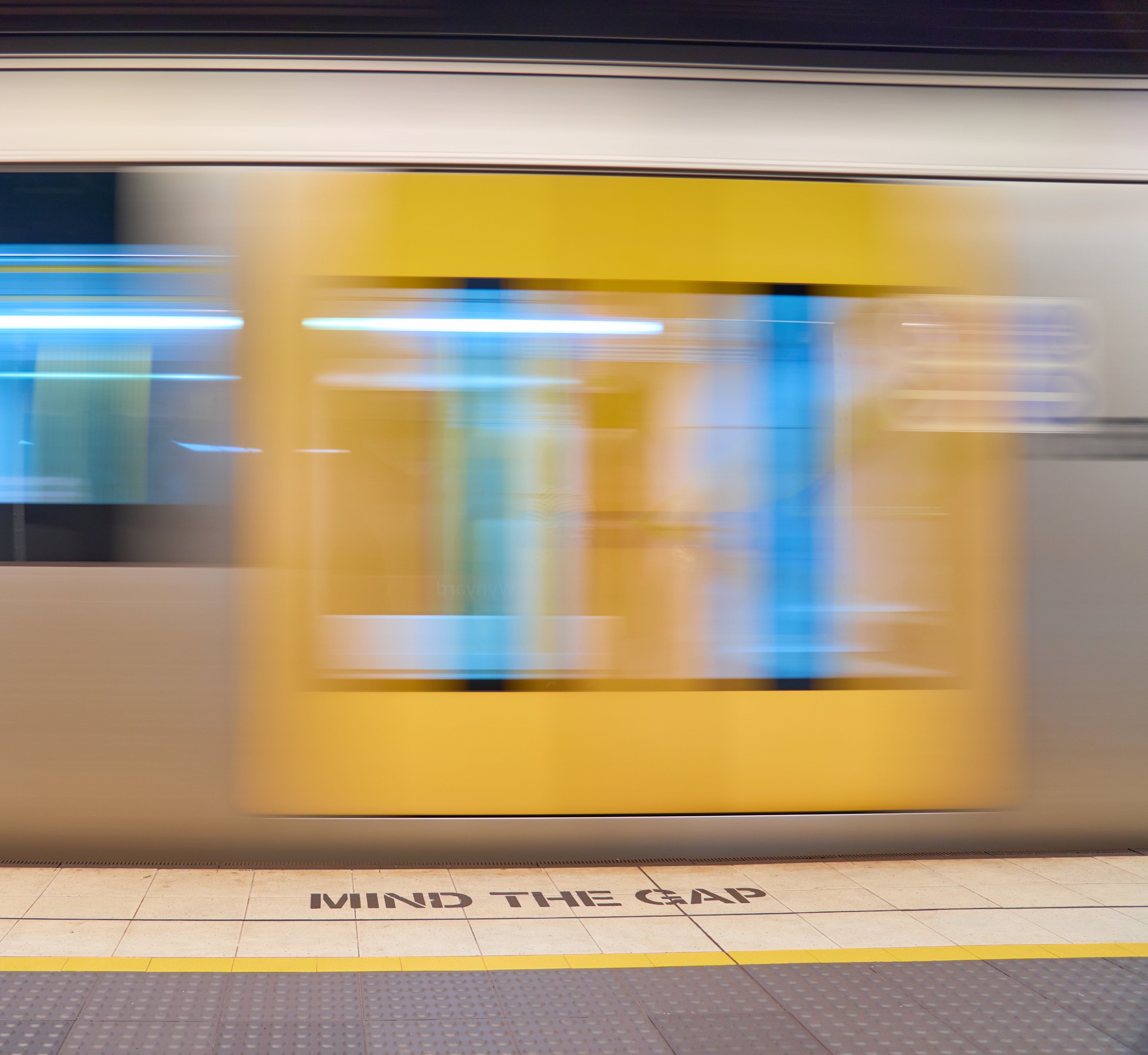Yellow subway train in motion at platform with 'MIND THE GAP' safety text visible on platform edge and dark tunnel ceiling.
