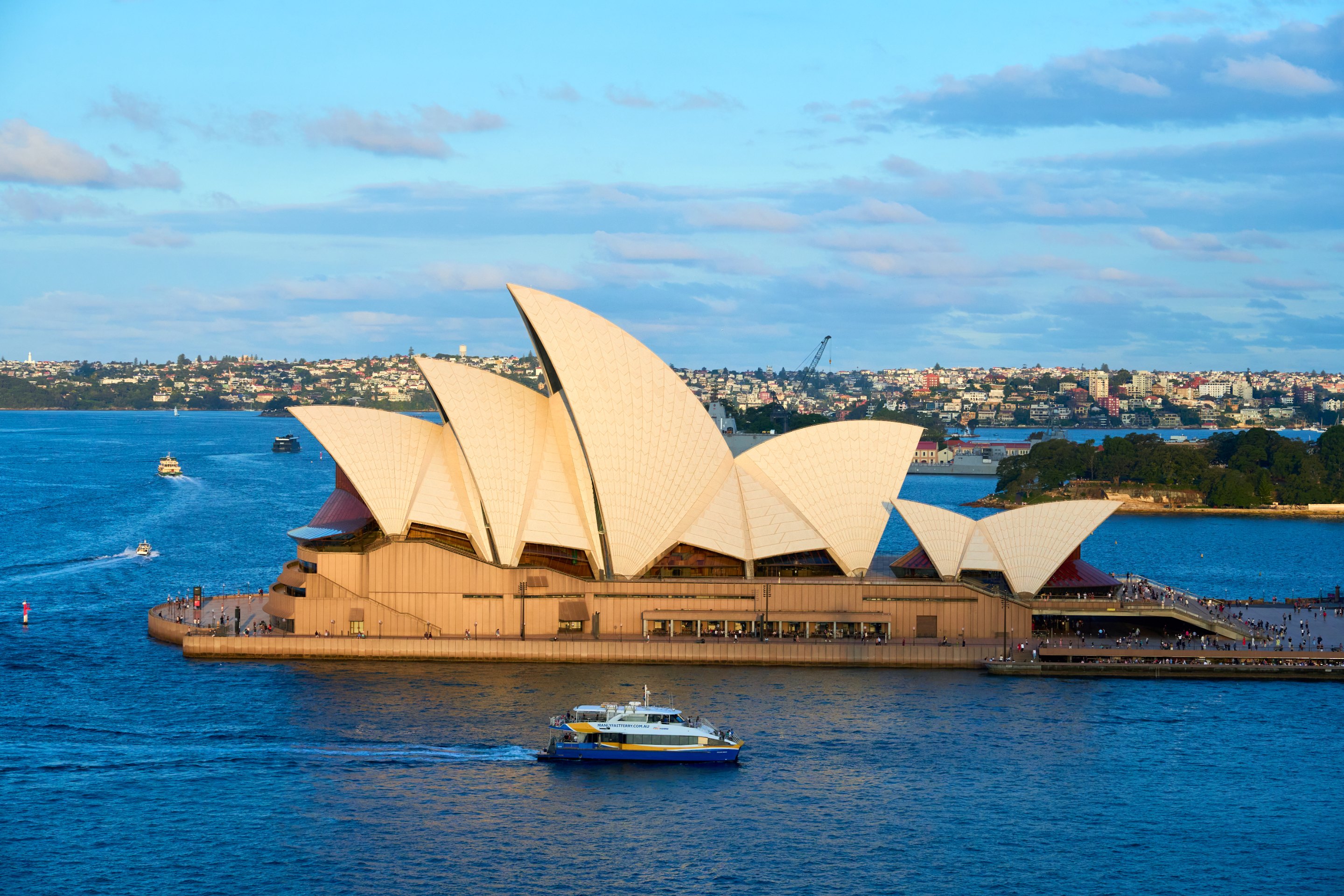 Sydney Opera House's white shell roofs lit gold at sunset, with a ferry crossing the harbour in front.