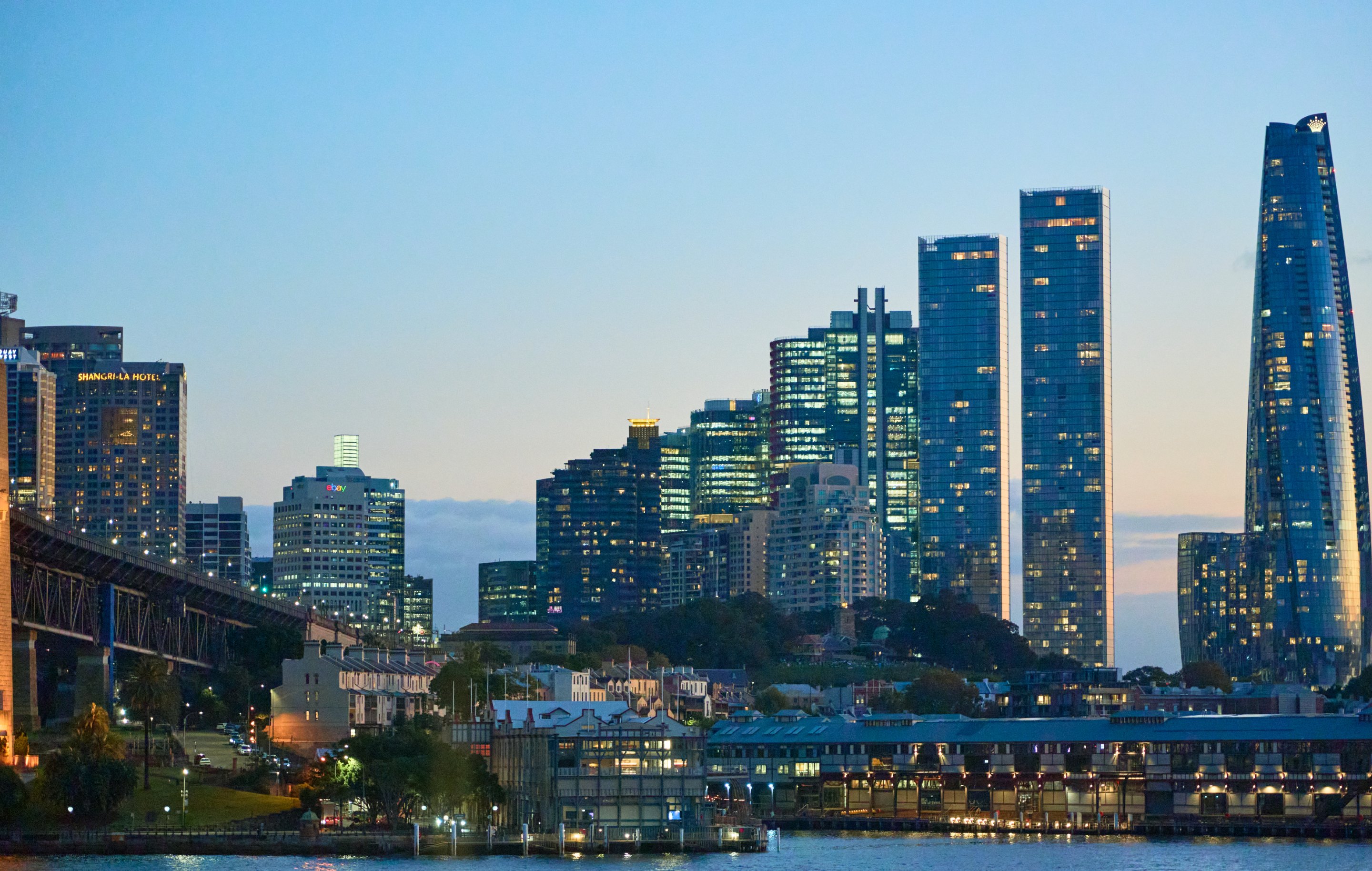 City skyline lit up at dusk, tall towers near the waterfront with a bridge and pier in the foreground.