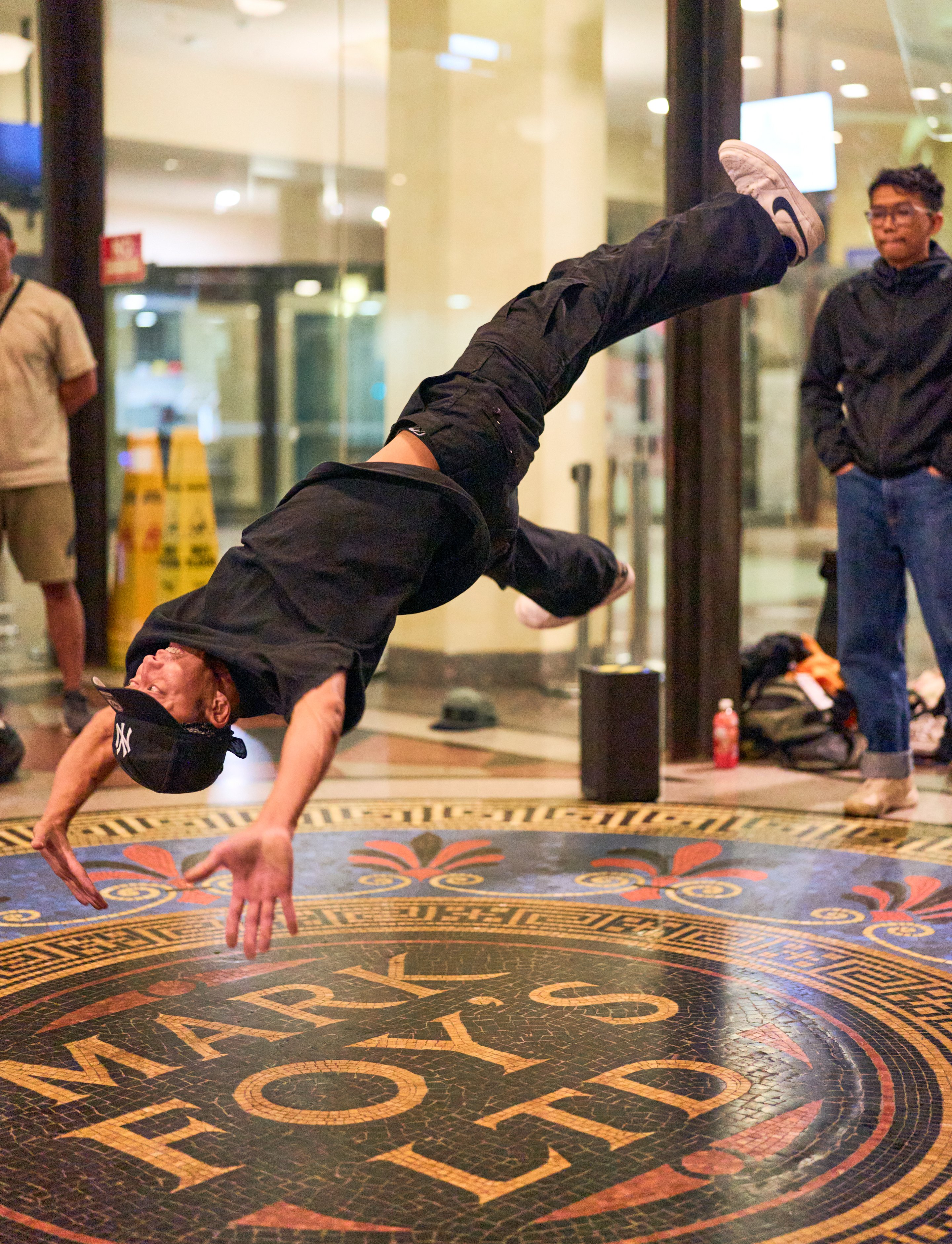 Person performing a headstand on an ornate patterned floor inside a modern building, with onlookers in the background.