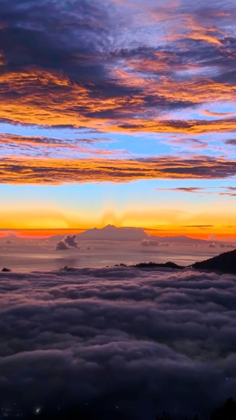 Sunrise or sunset view from above cloud cover showing a mountain in silhouette against a dramatic orange, purple, and blue sky.