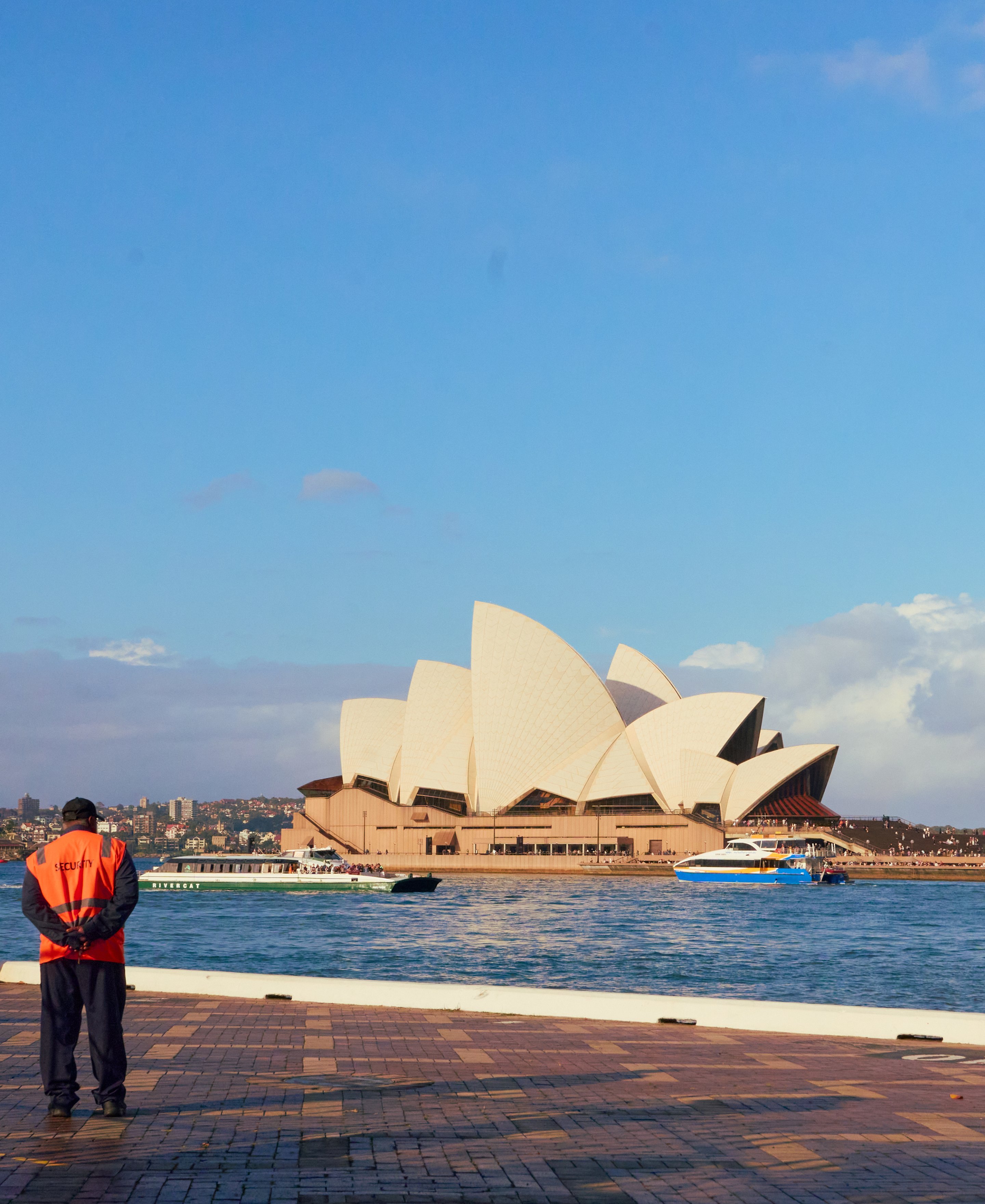 Person in orange safety vest standing on waterfront promenade facing Sydney Opera House across blue harbor water.