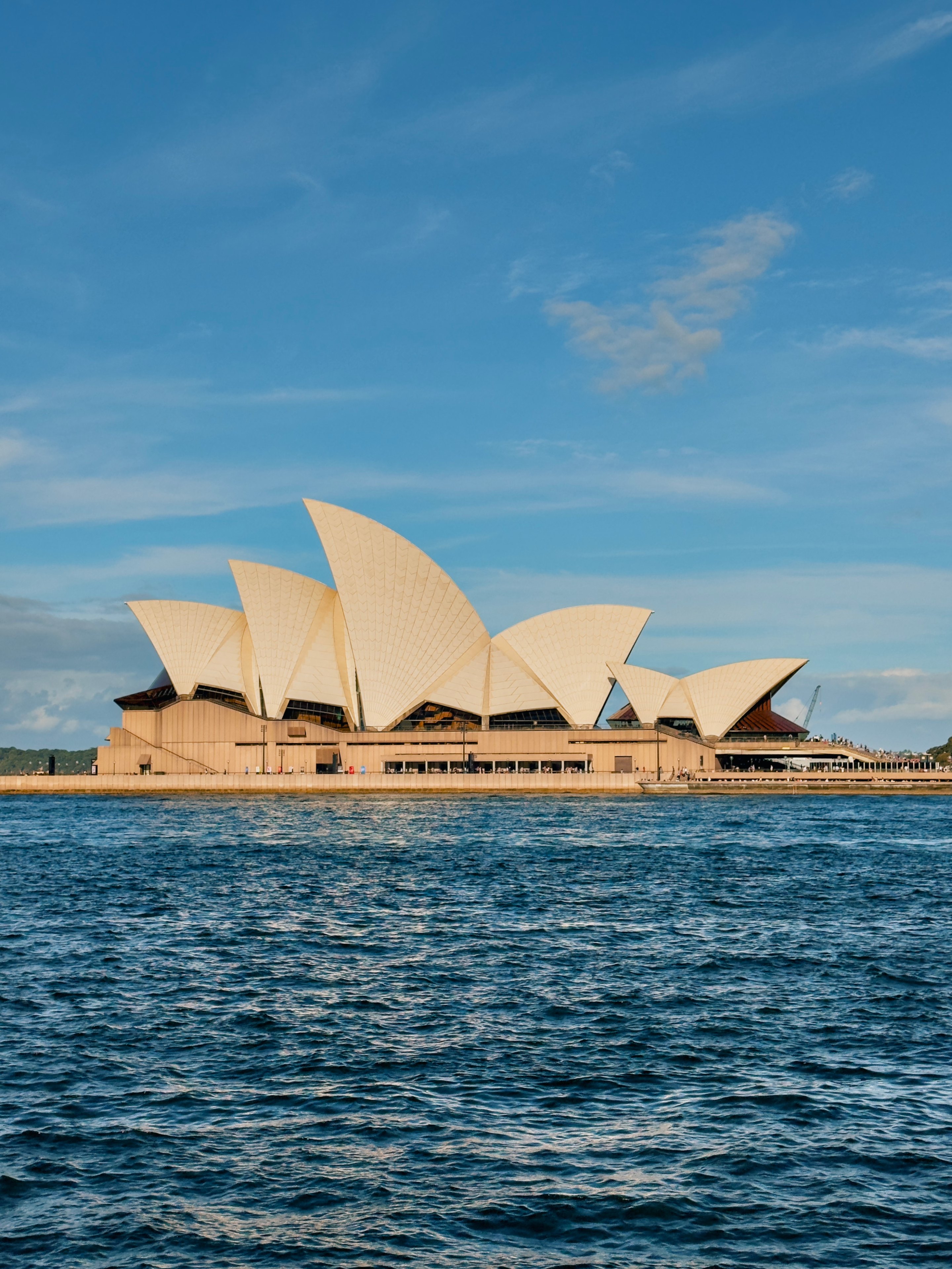 Sydney Opera House viewed from harbor with distinctive white shell-shaped structures against clear blue sky and blue water.