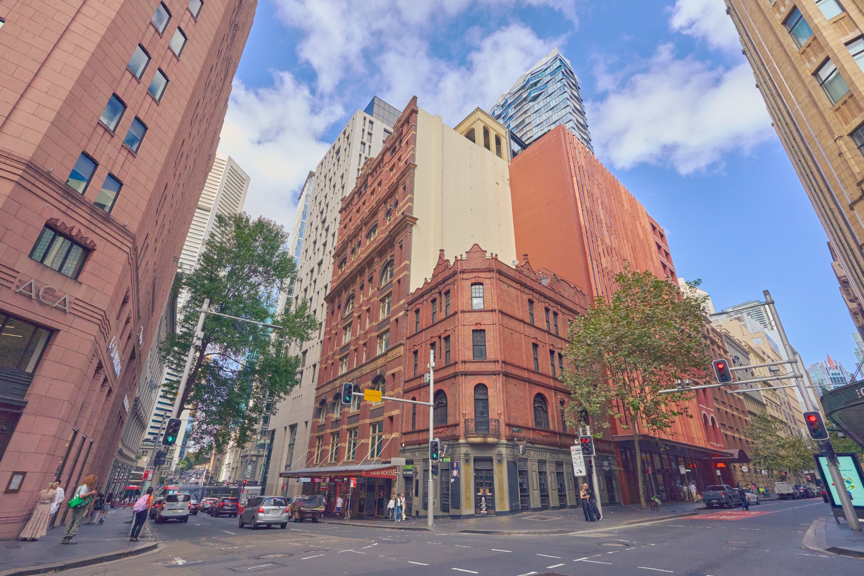 Historic urban street with ornate red-brick buildings of varied heights, mature street trees, and clear blue sky.