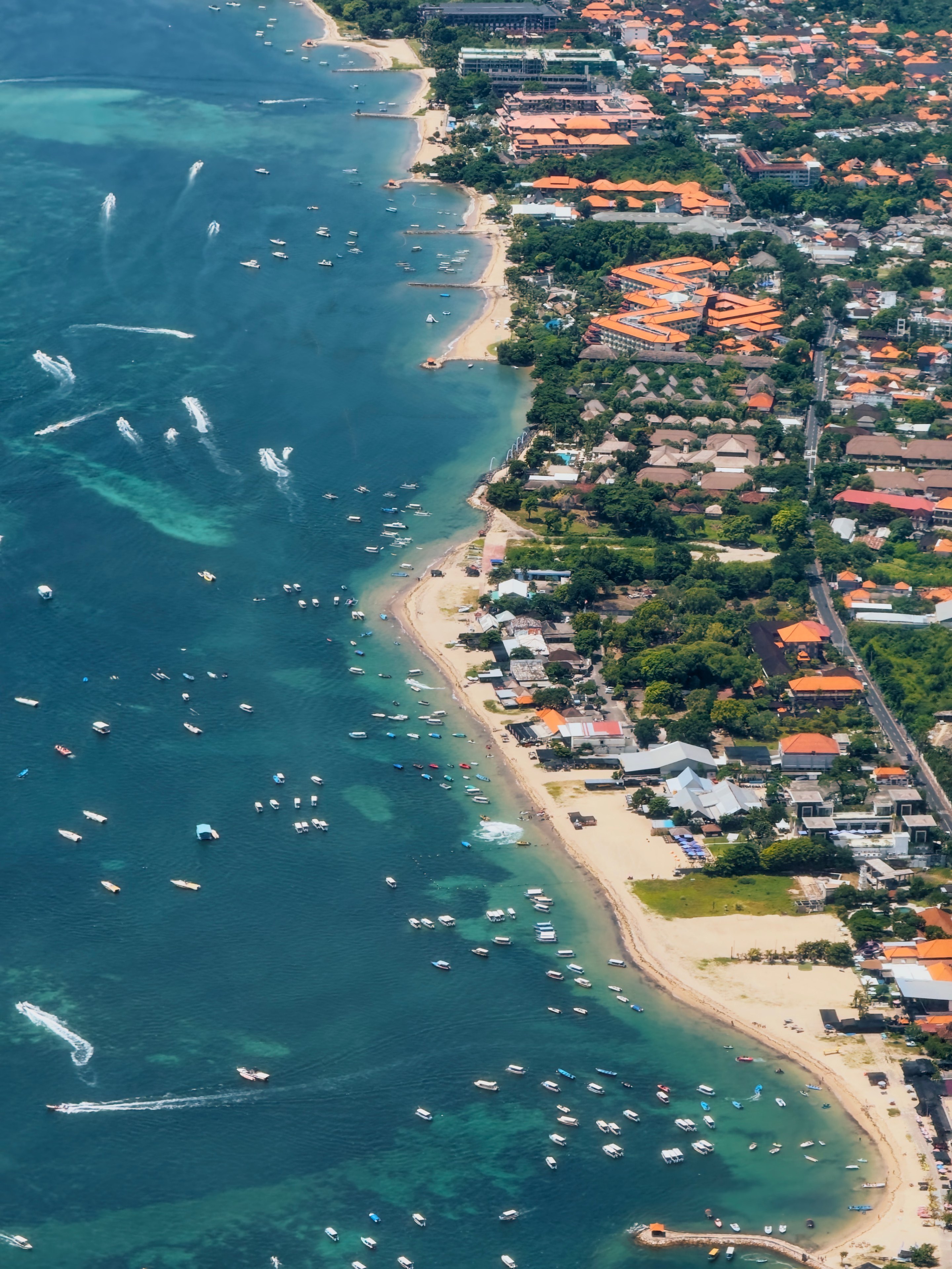 Aerial view of coastal town with turquoise ocean dotted with white sailboats, sandy beach, orange-roofed buildings, and green vegetation along shoreline.