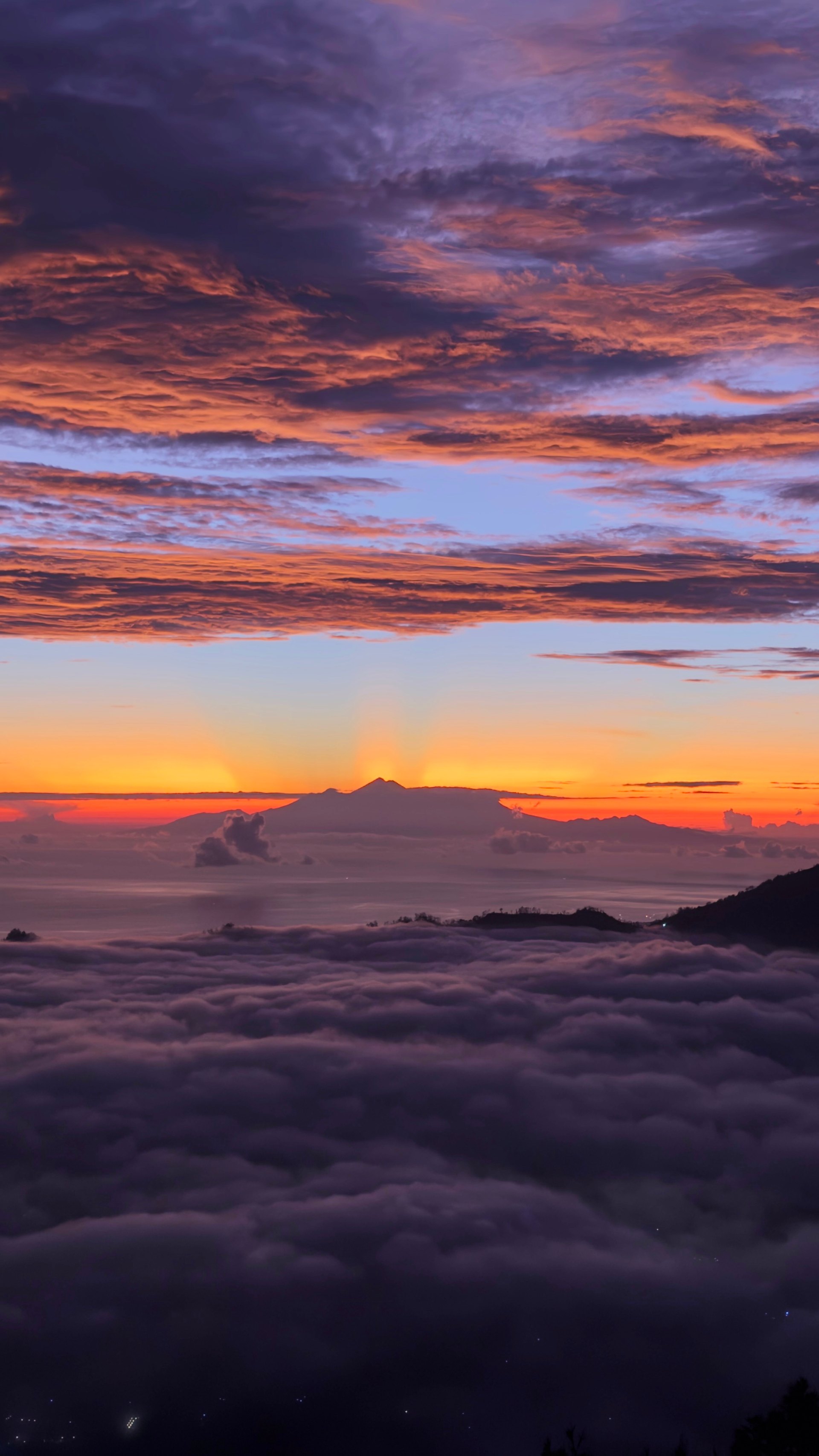 Sunrise over layered clouds and misty seascape with island silhouettes on horizon and sun glowing at water line, warm orange and purple tones filling sky.