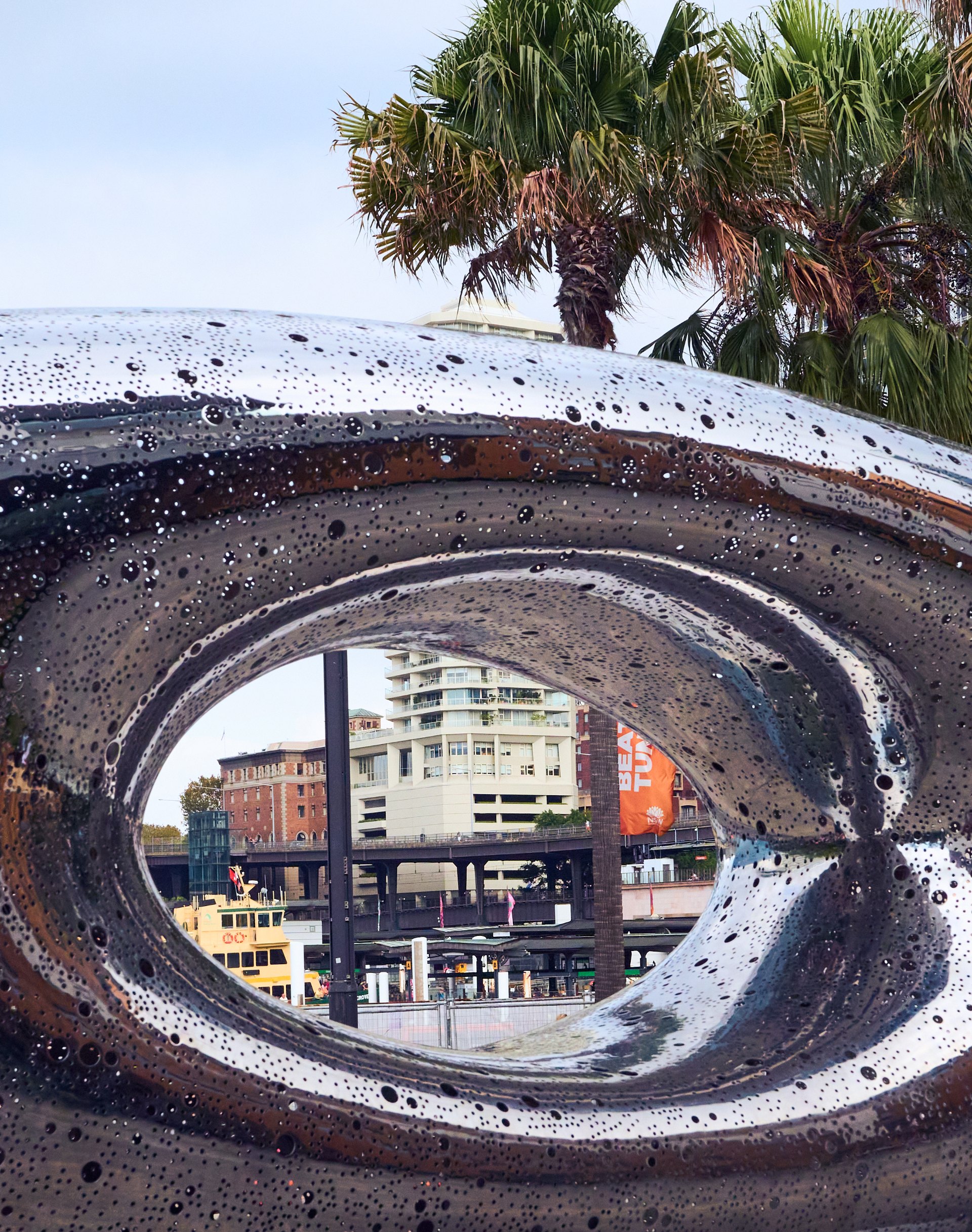 Weathered circular metal pipe or structure framing view of beachfront buildings and palm trees, seafoam spray visible on pipe edges.