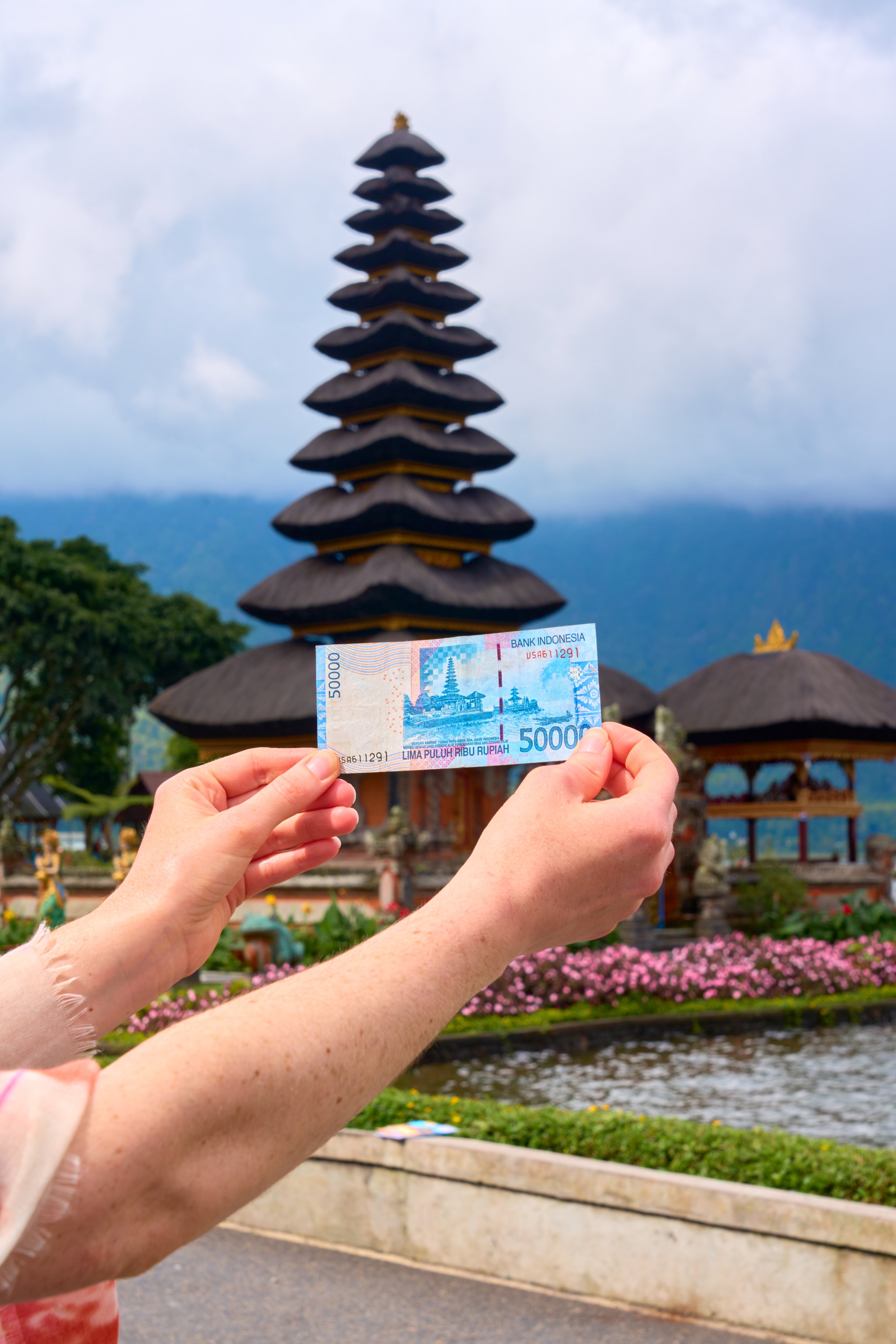 Hand holding an Indonesian banknote featuring a temple image, photographed in front of the actual tiered temple structure with mountains or ocean in the distance.