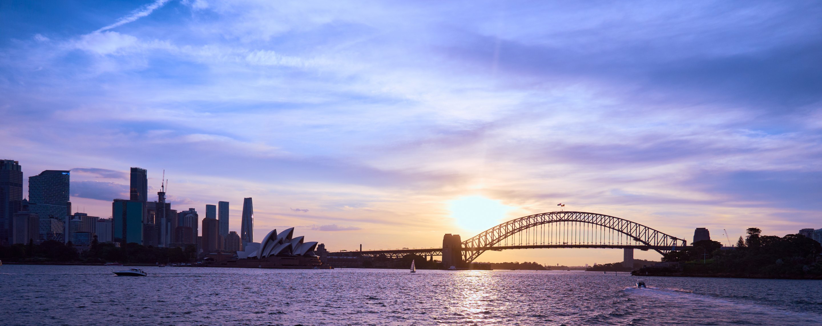 Sydney Harbour Bridge's distinctive arch silhouetted at sunset with city skyline, calm water, and purple-tinted sky.