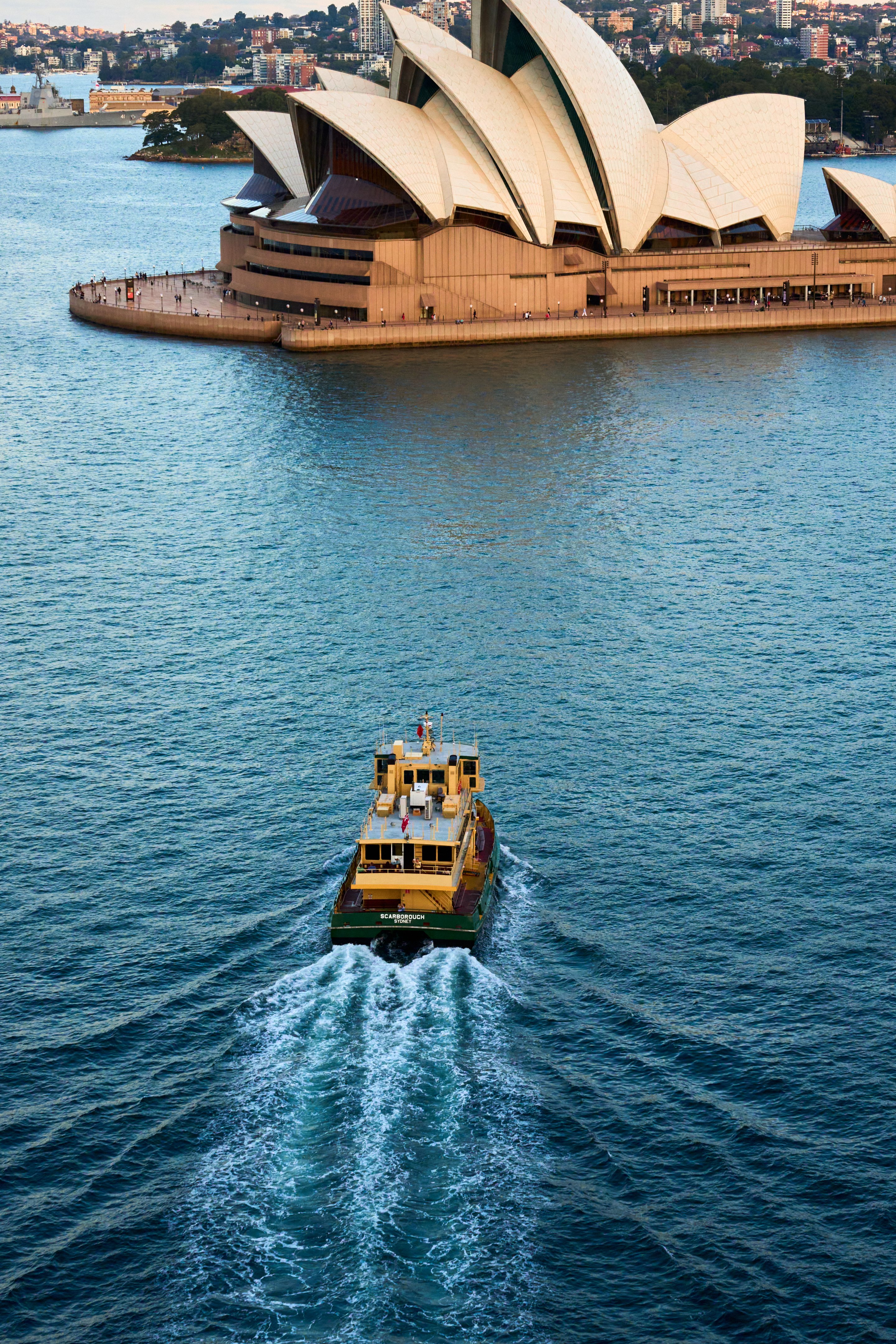 Elevated view of a Sydney ferry approaching the Opera House, its wake trailing across the harbour.