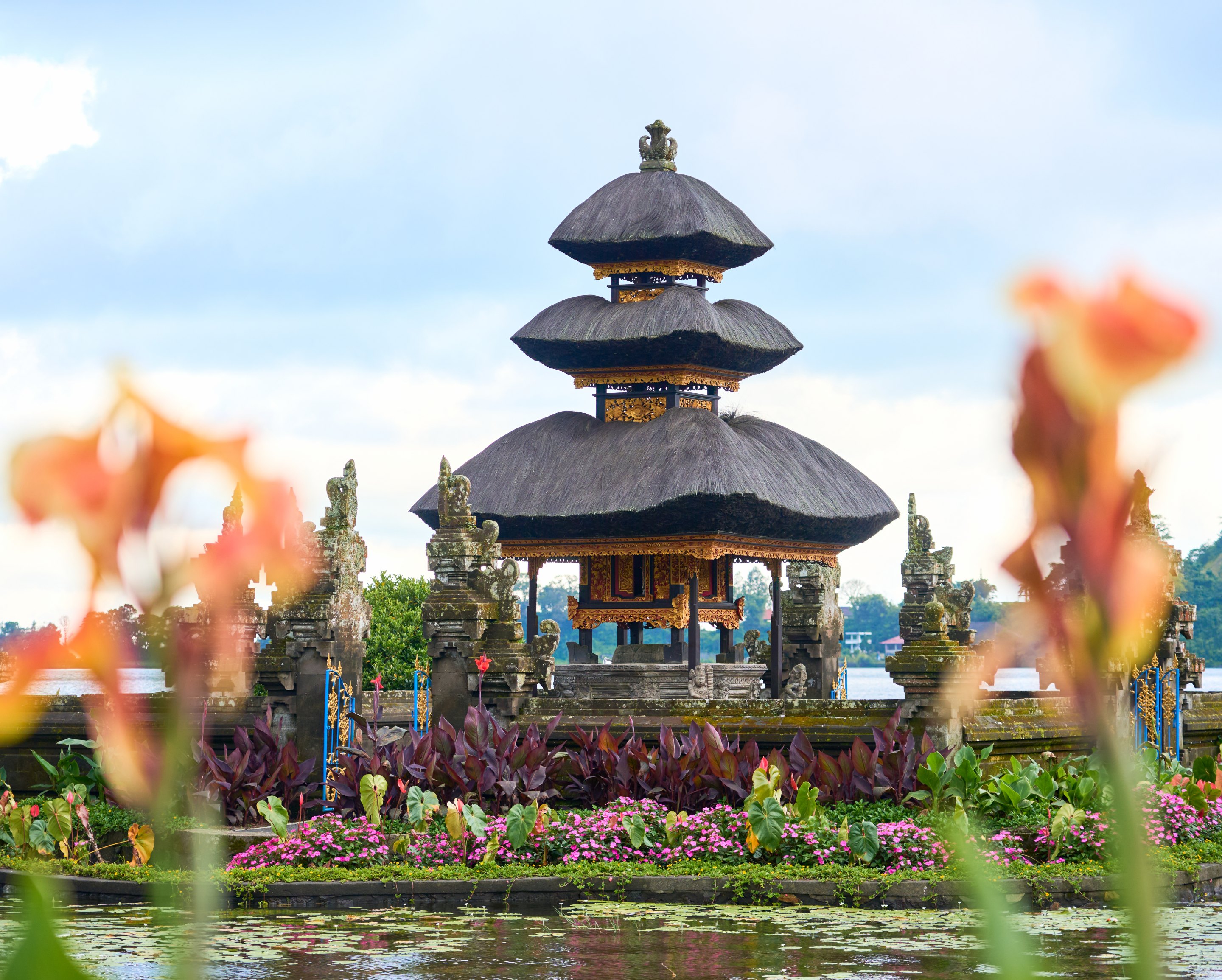 Balinese temple with tiered roofs framed through blooming flowers in the foreground, with additional temple structures and landscaping visible.