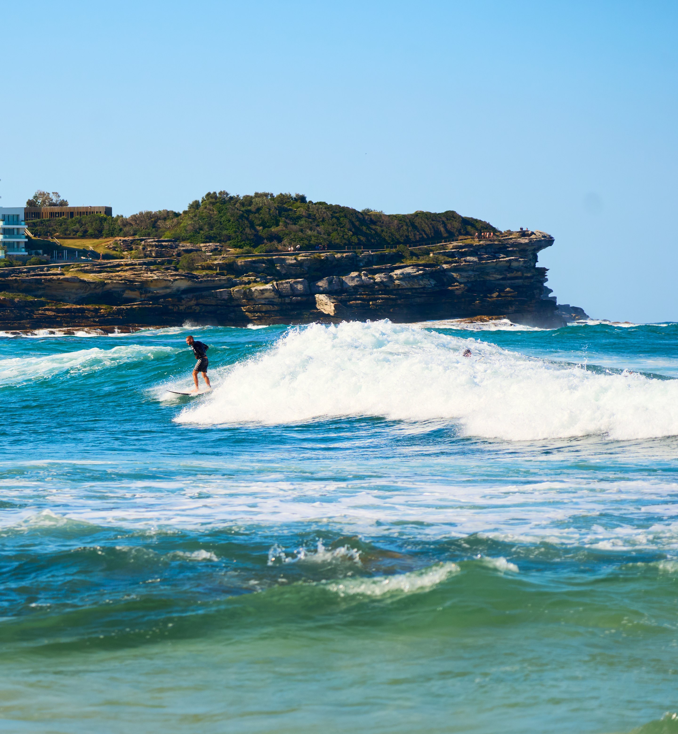 A surfer rides a white wave in turquoise water, with a rocky coastal headland thick with vegetation rising in the background beneath a clear blue sky.