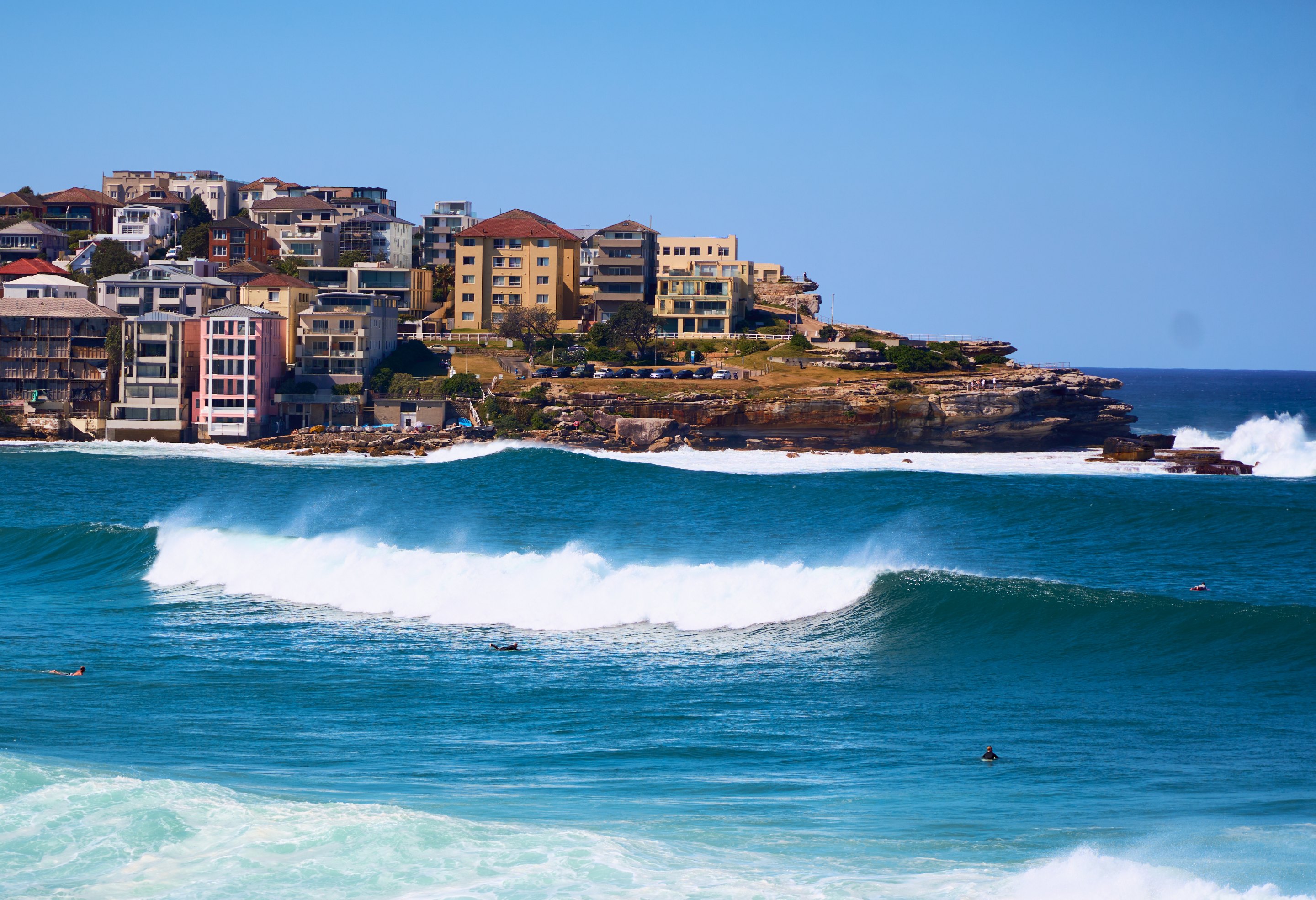 Apartment buildings crowding a coastal headland overlooking turquoise water and a breaking wave, with a swimmer visible in the water.