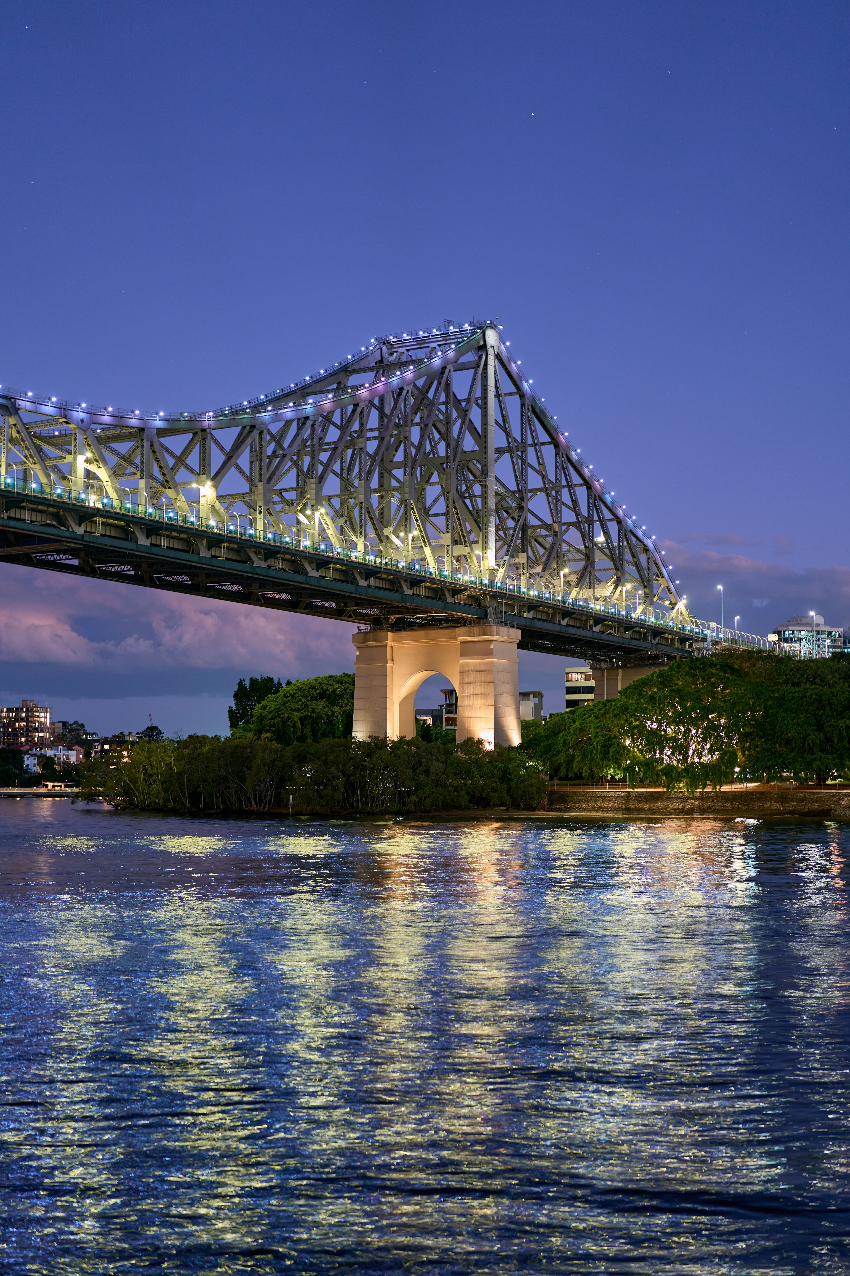 The Story Bridge lit with white lights at twilight, reflecting in the water below, with trees and city buildings underneath and deep blue evening sky above.