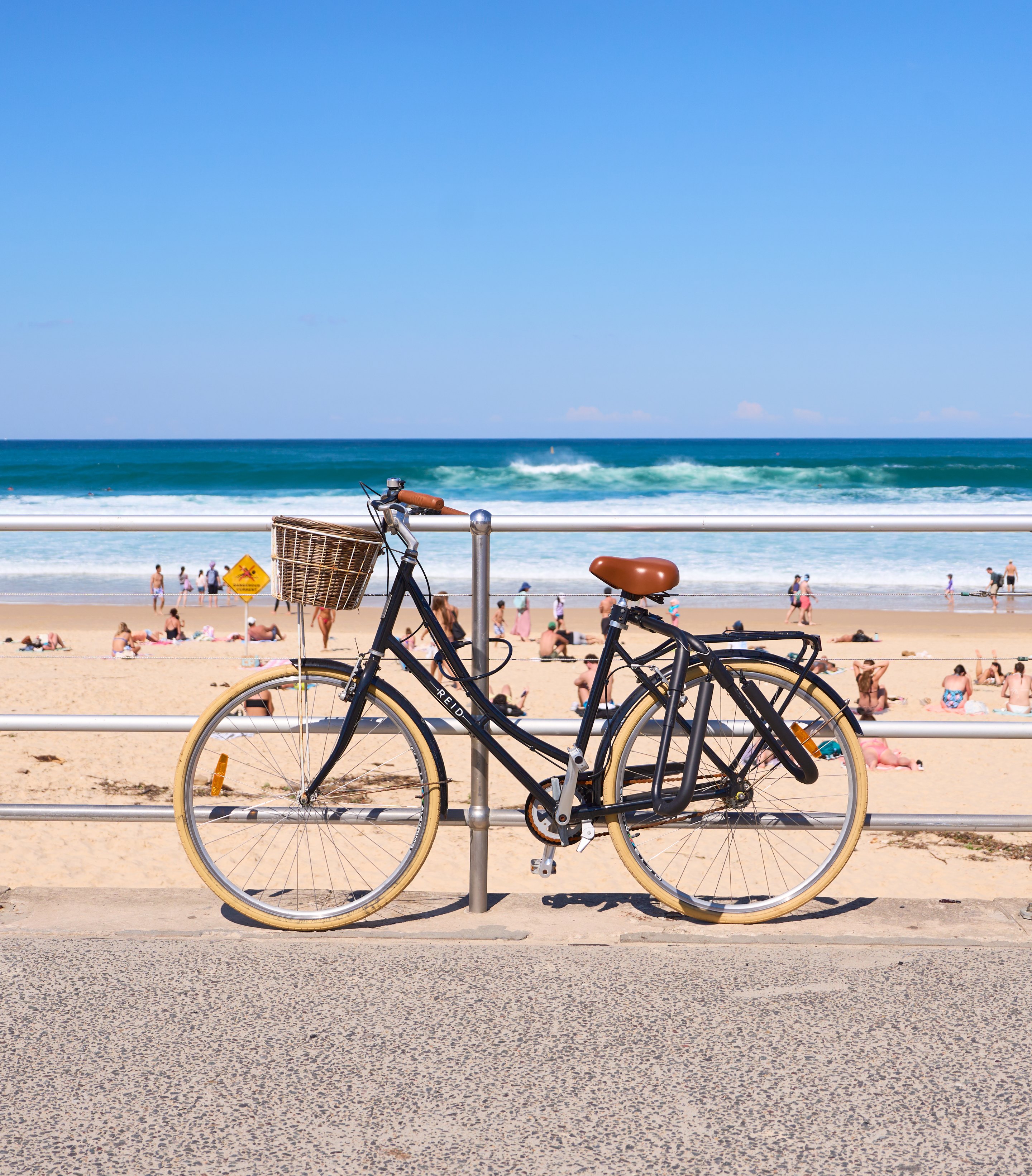 Black bicycle parked on a sandy beach promenade overlooking swimmers and sunbathers in turquoise water.