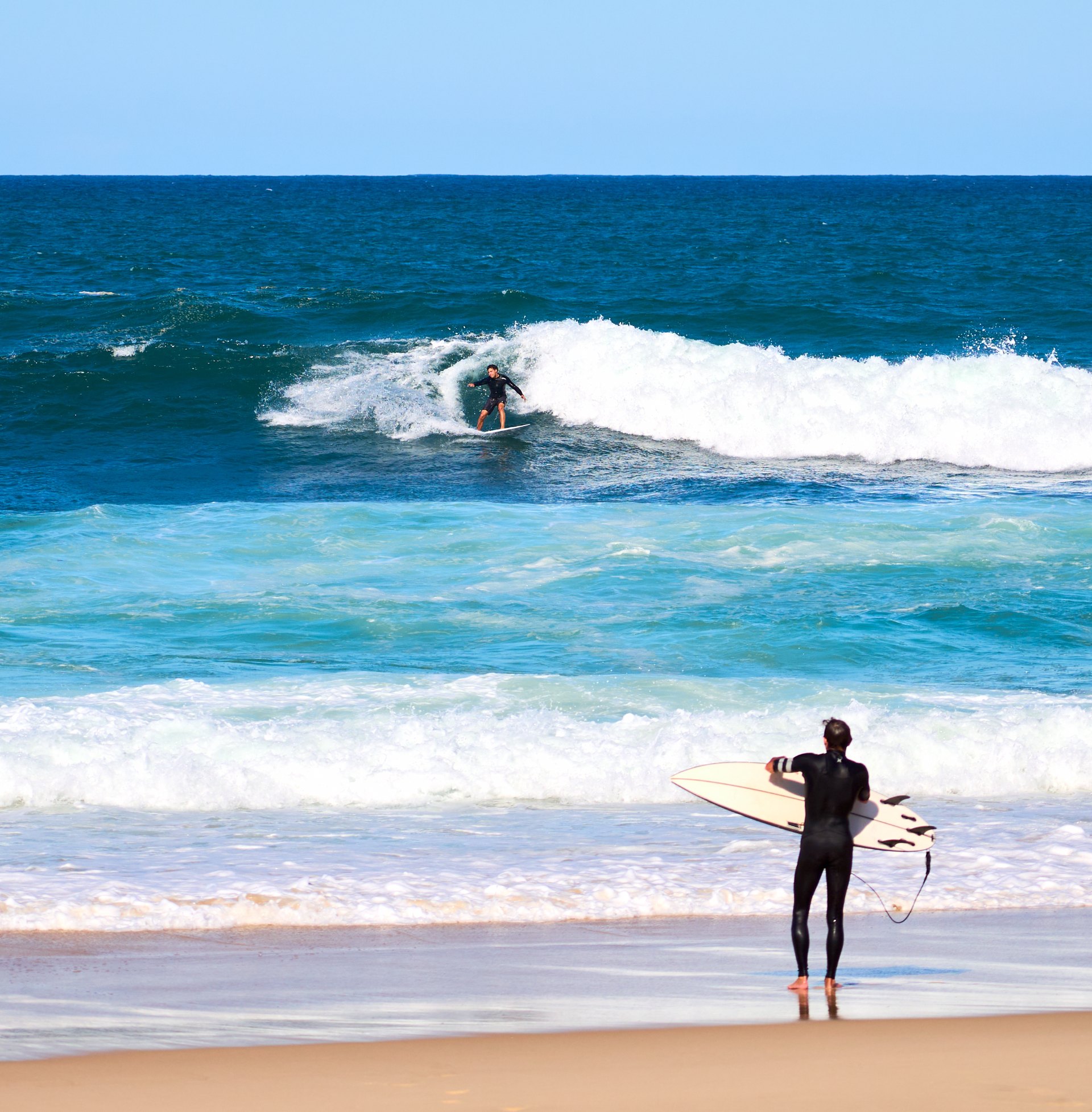 Surfer in dark wetsuit standing on the beach holding a white surfboard, watching another surfer ride a distant wave.