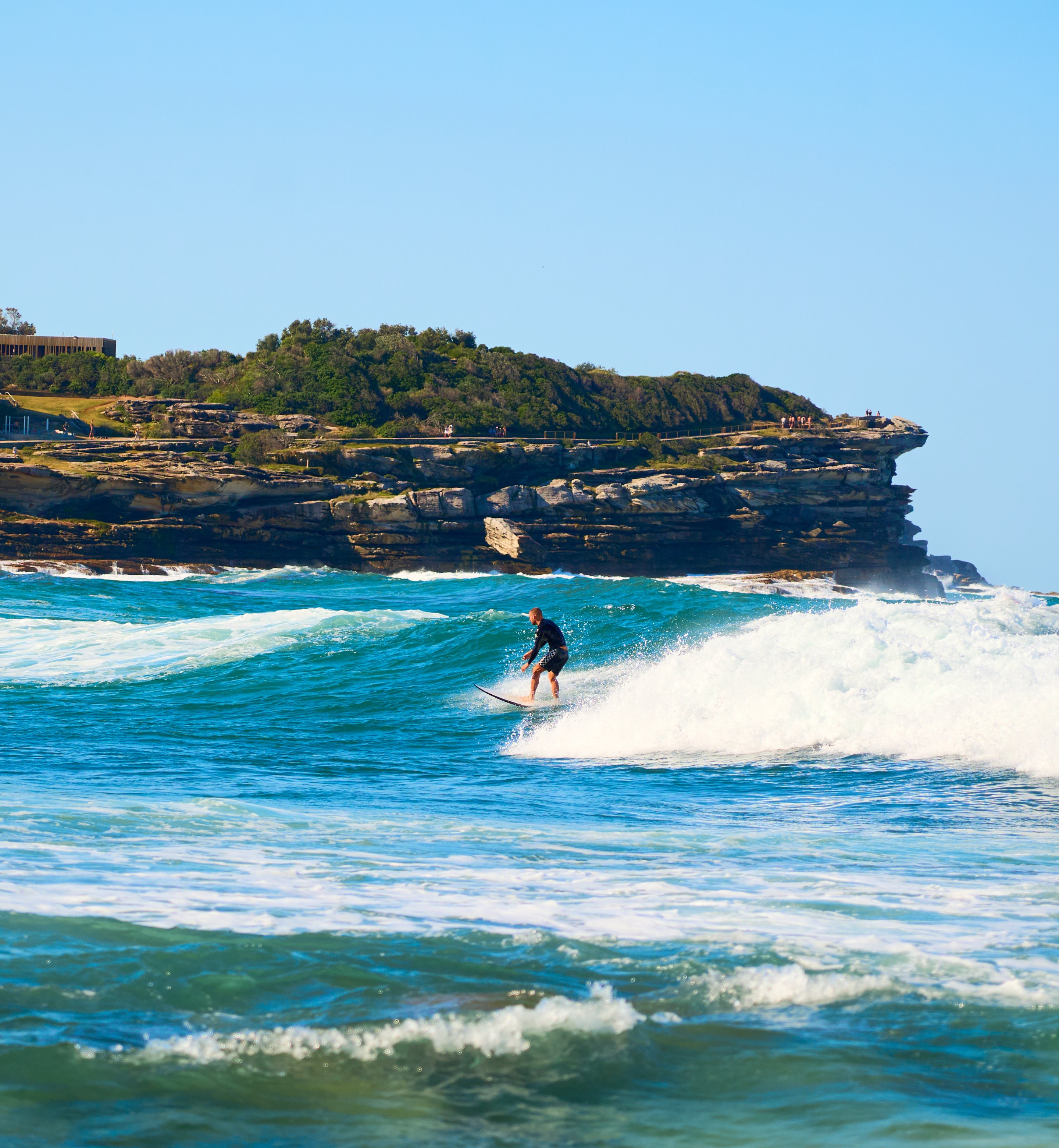 Surfer riding a wave with a dramatically layered rock headland and green vegetation towering in the background.