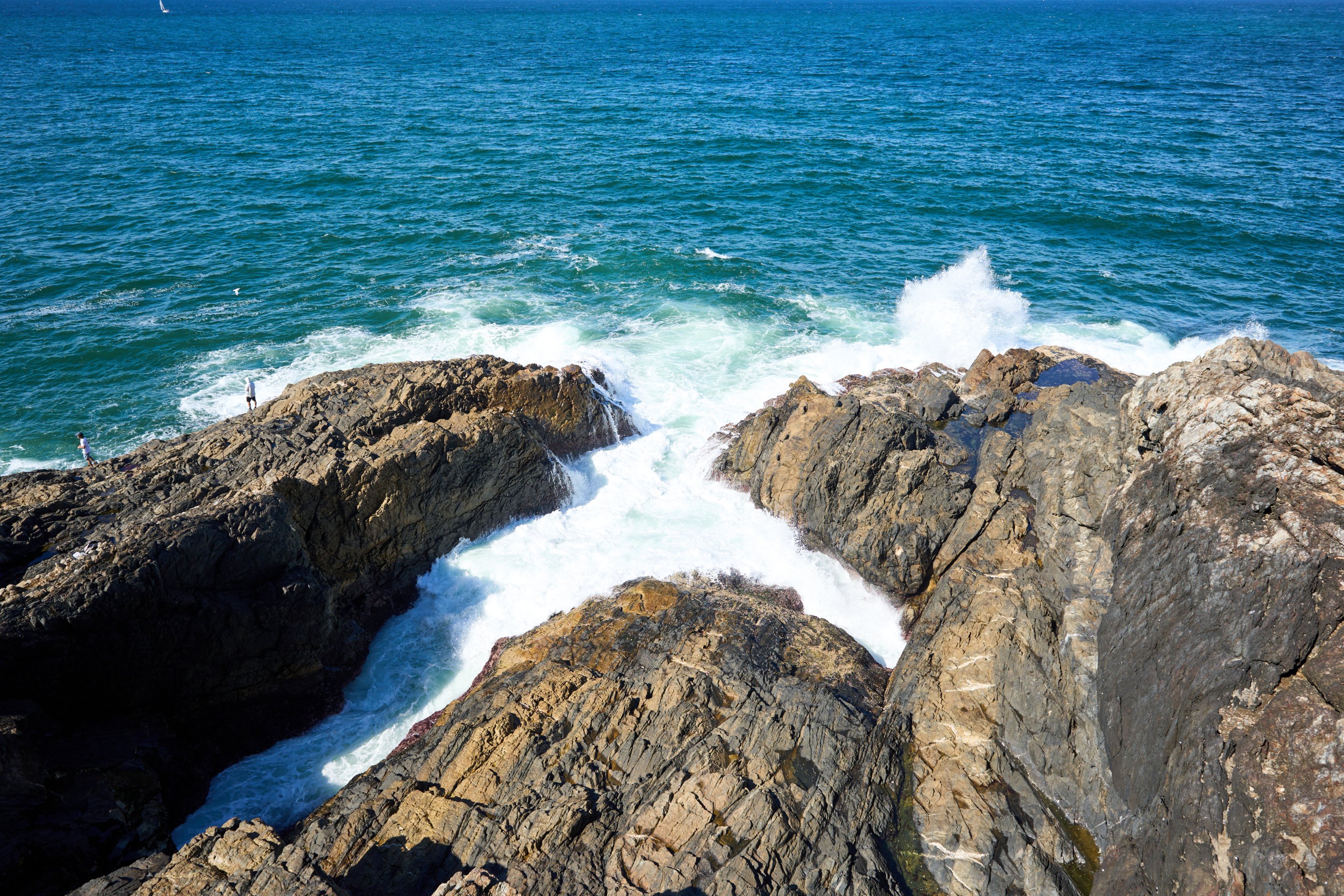 Overhead view of waves crashing in white spray against layered dark rock formations on a headland, with blue-green water beyond.