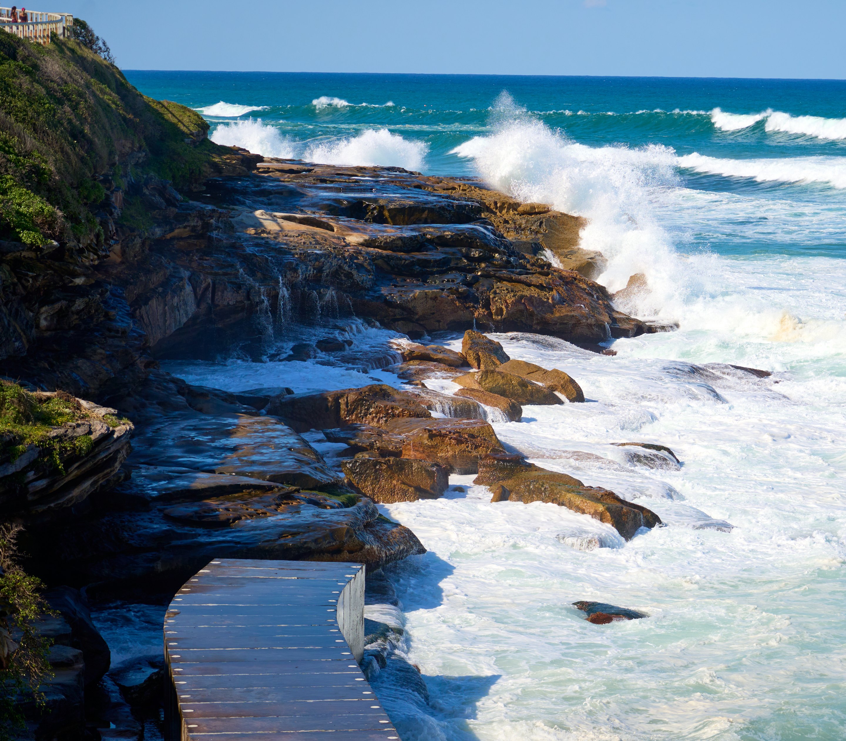 Dark, jagged rock formations with white waves crashing against them, green cliff vegetation visible above.