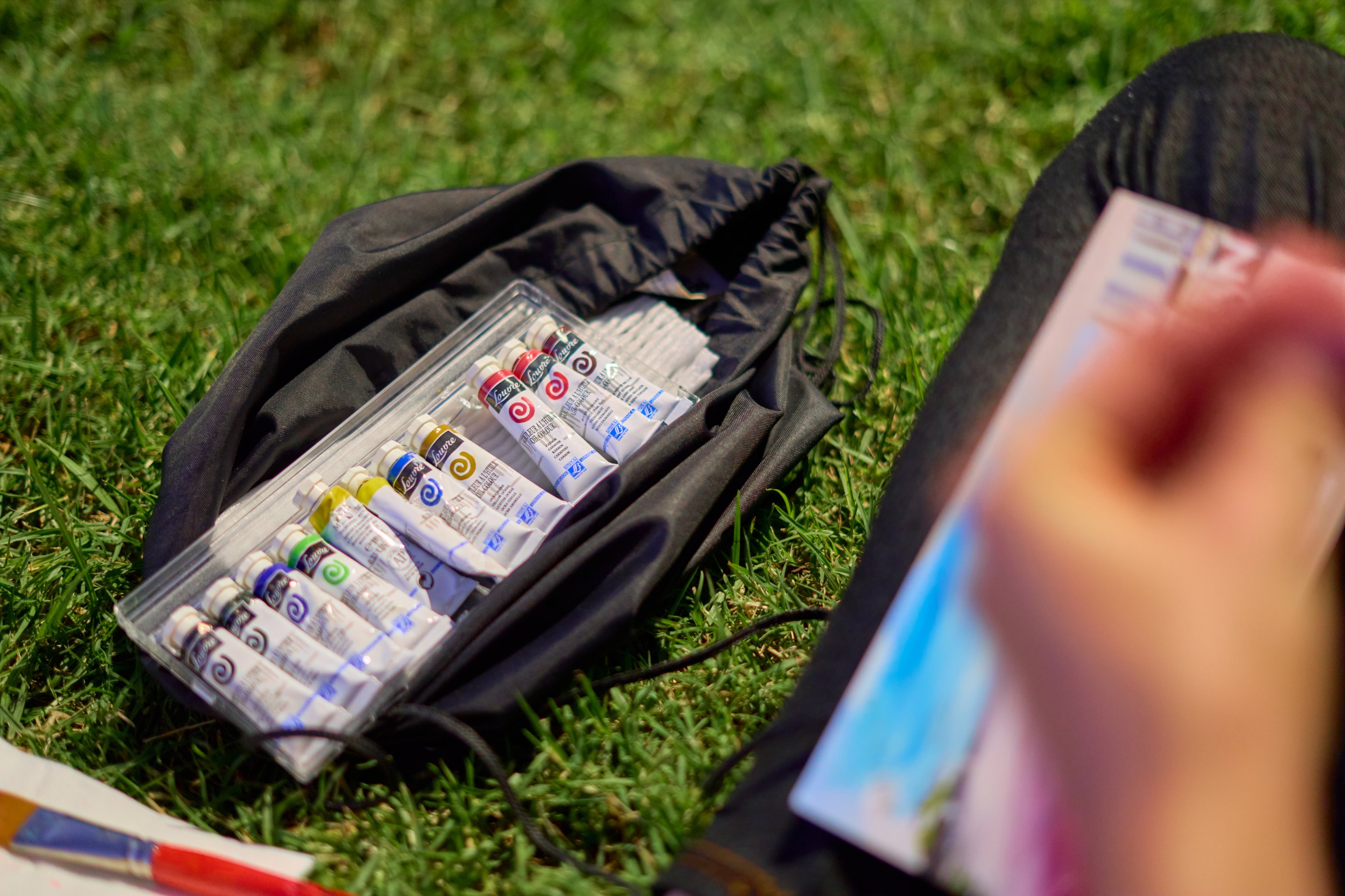 A set of watercolour paint tubes laid out on grass, with a hand holding a small painted card nearby.