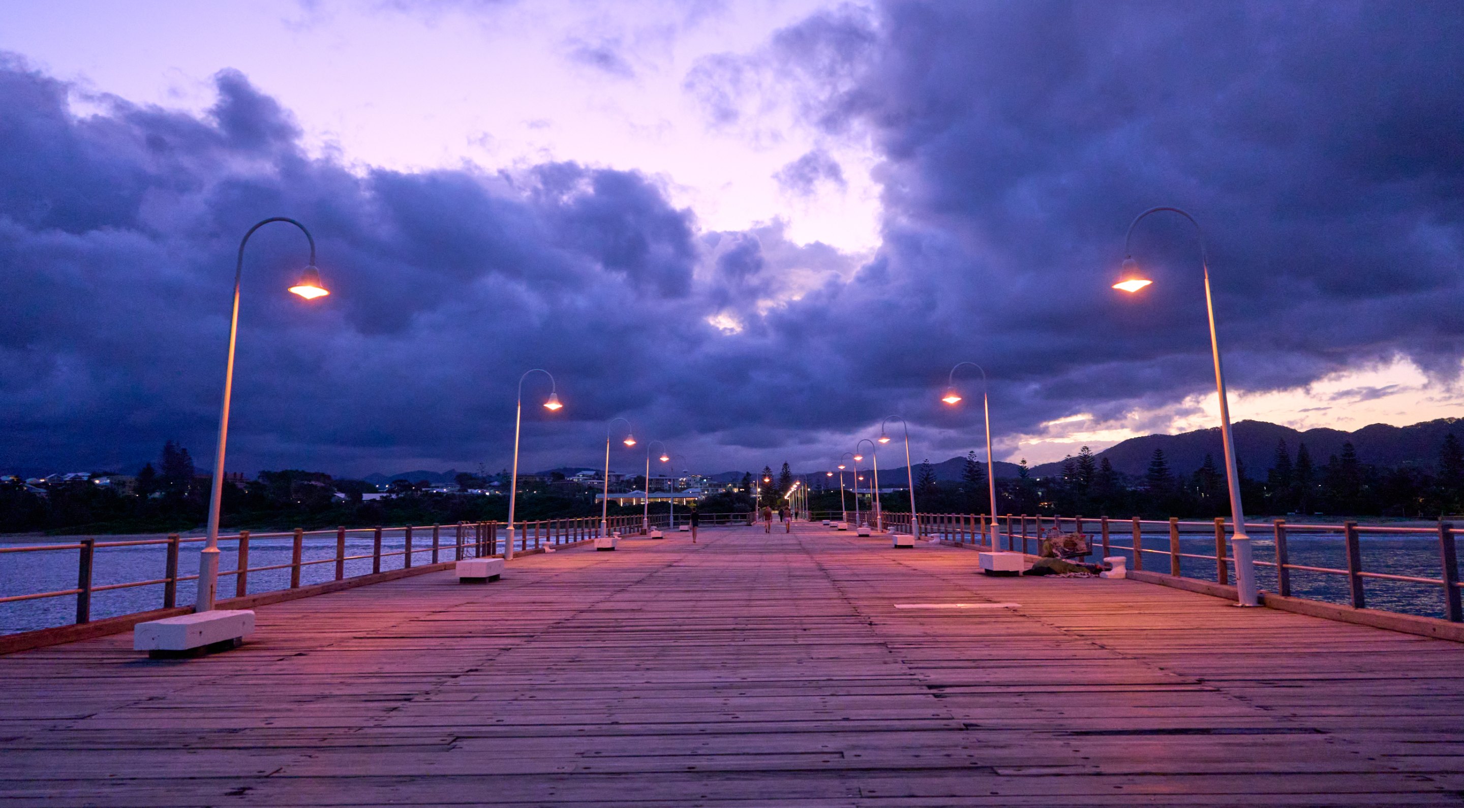 A wooden pier stretches empty toward the water at twilight, lit by overhead street lamps, with purple-pink clouds and blue sky above.
