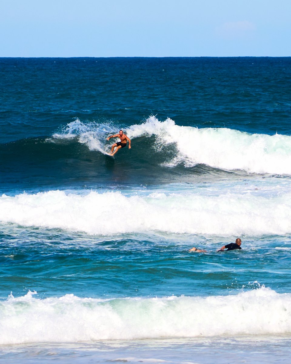 Surfer in bright orange top riding inside the white foam of a breaking wave, another swimmer visible in background.