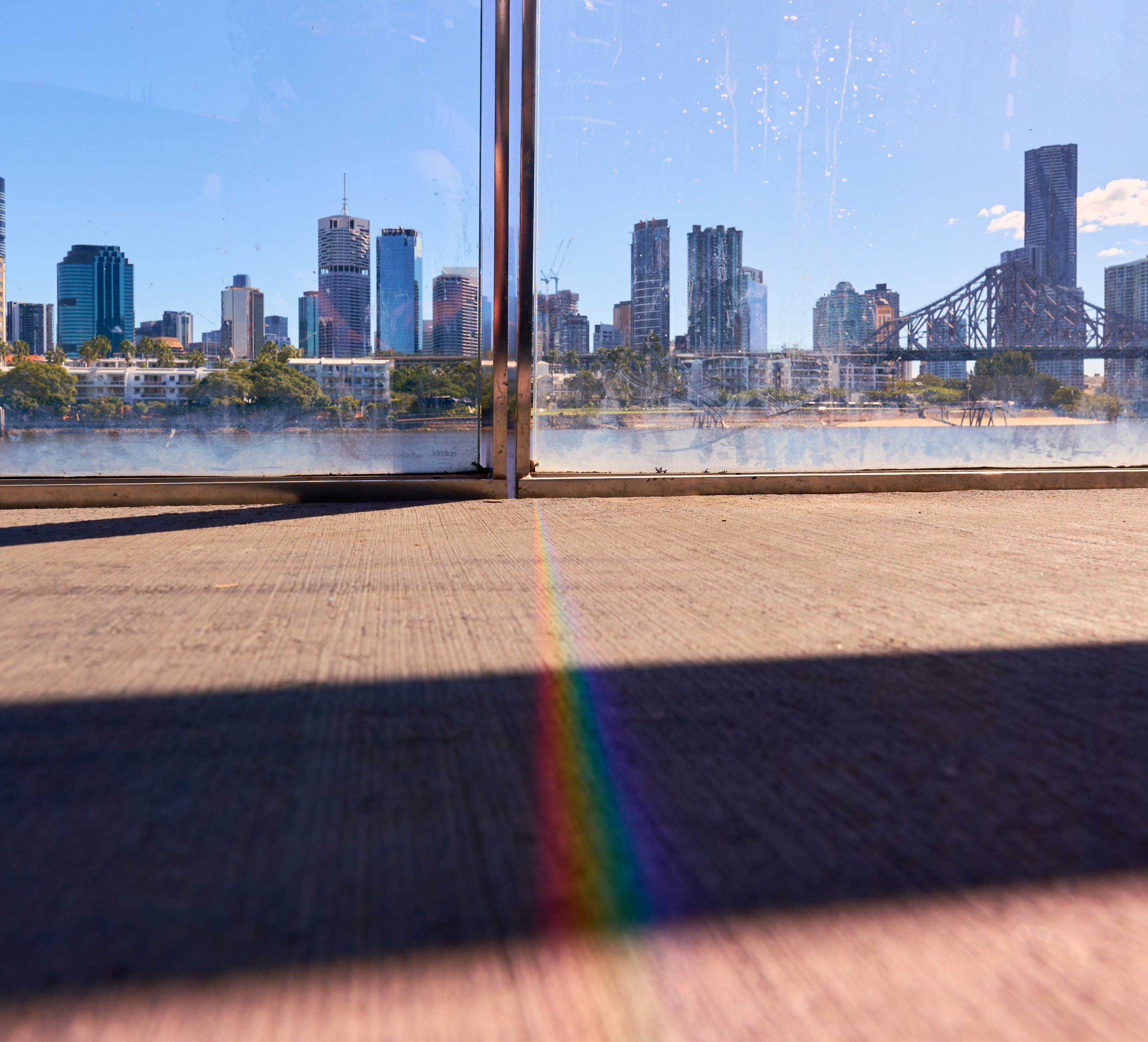 City skyline viewed through a window with a brown metal support beam dividing the frame, river below reflecting light, wooden boardwalk in foreground, and rainbow prism in lower corner.