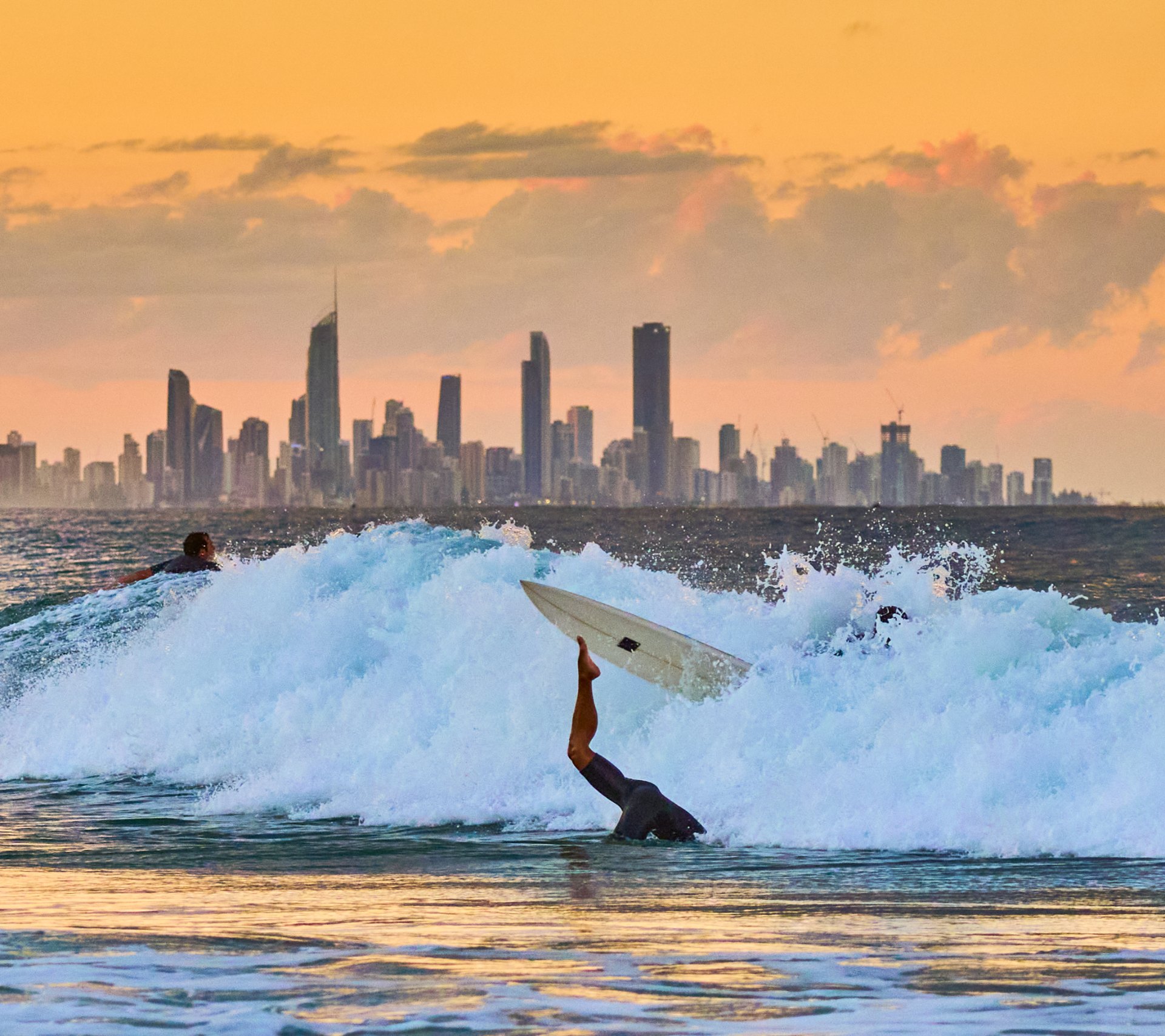 Surfer wiping out as their board flies out of the water, city skyline visible on the horizon at sunset.