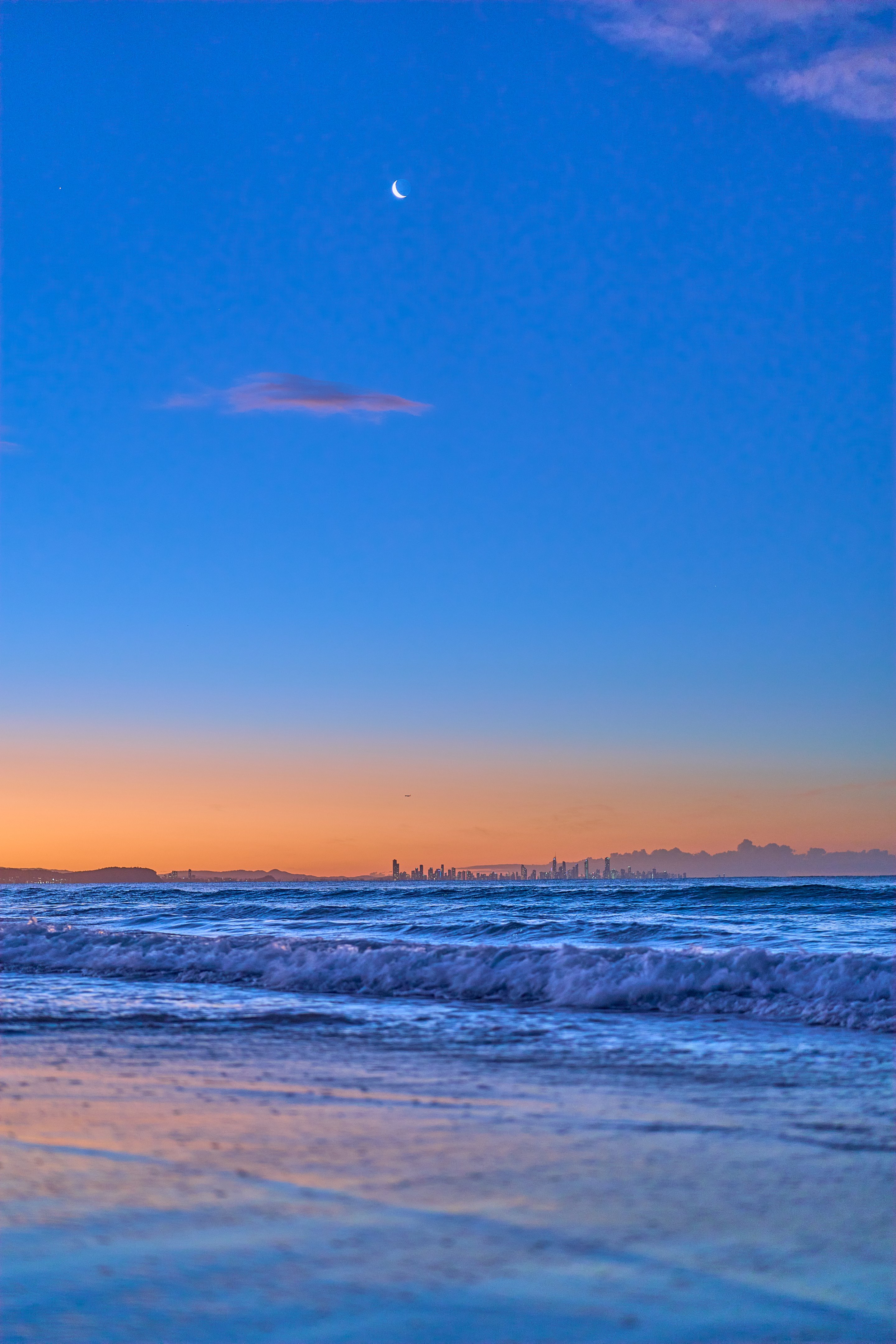Beach at sunset with waves, wet sand, and city skyline on the horizon, crescent moon visible in the blue sky above.