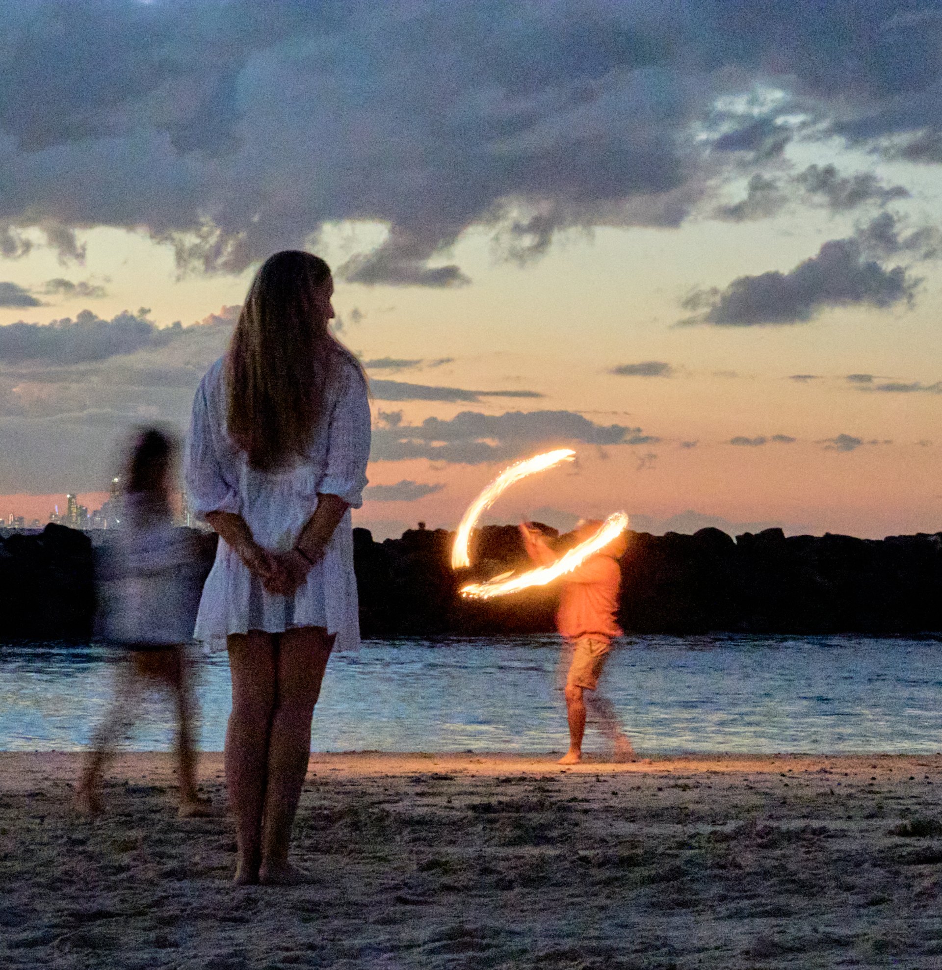 A woman stands on a beach at dusk while a child walks nearby and another person swings a glowing light, leaving a bright trail in the air.