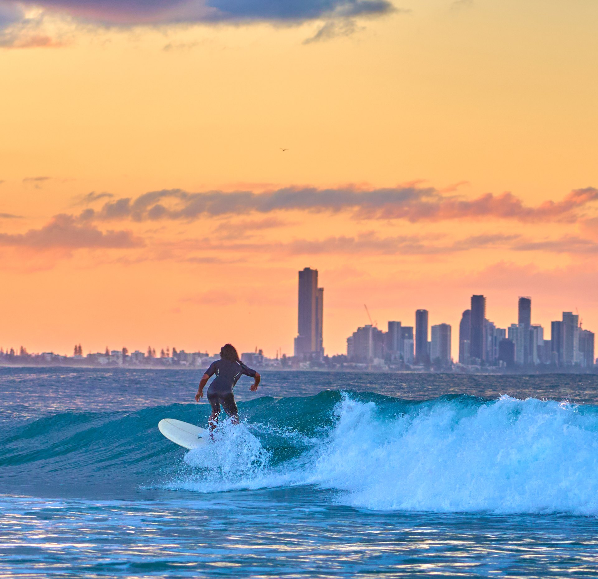 Surfer riding a wave at sunset with a city skyline silhouetted on the horizon.