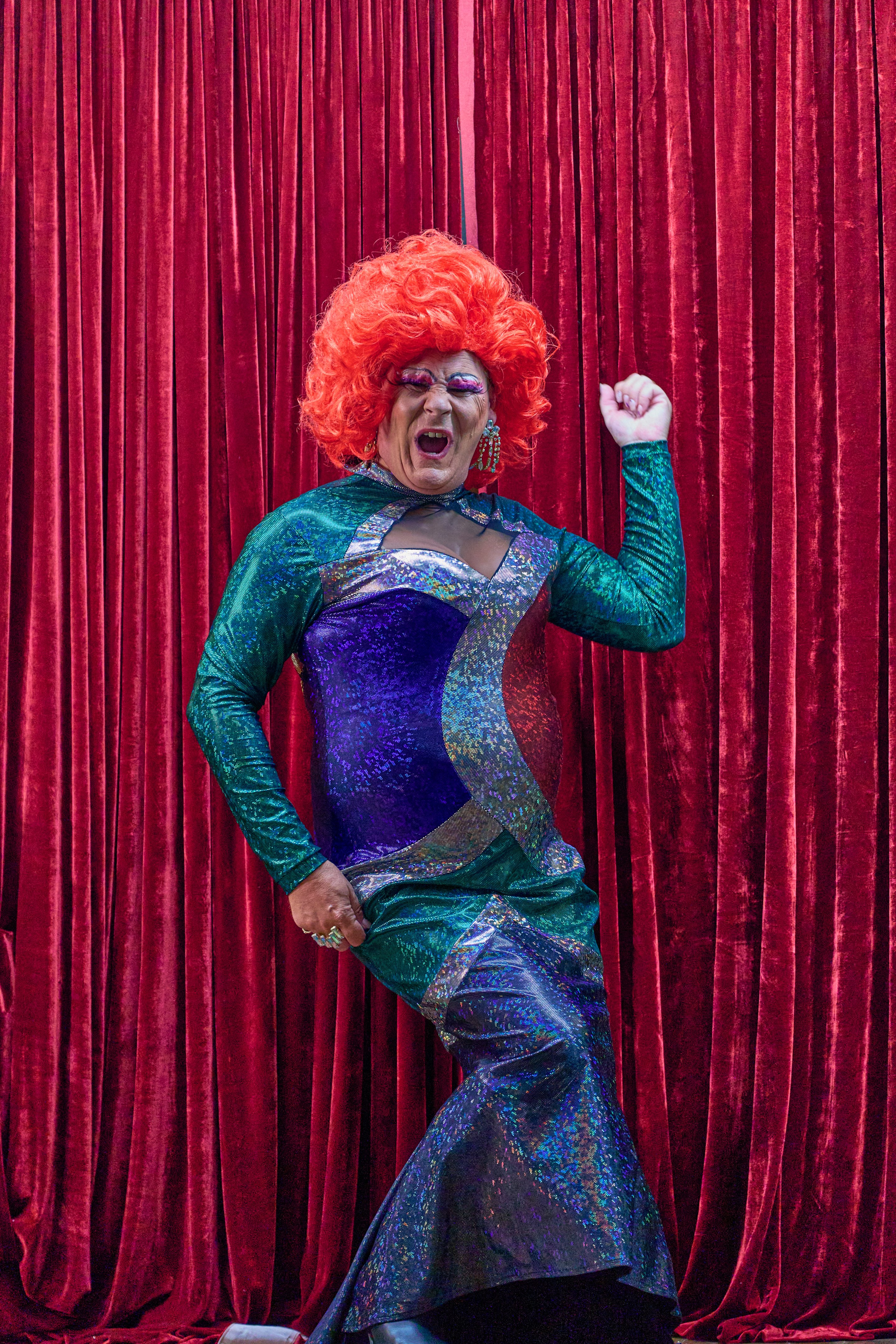 Performer in orange afro wig and sparkly blue sequined outfit strikes a triumphant pose against deep red velvet curtains.