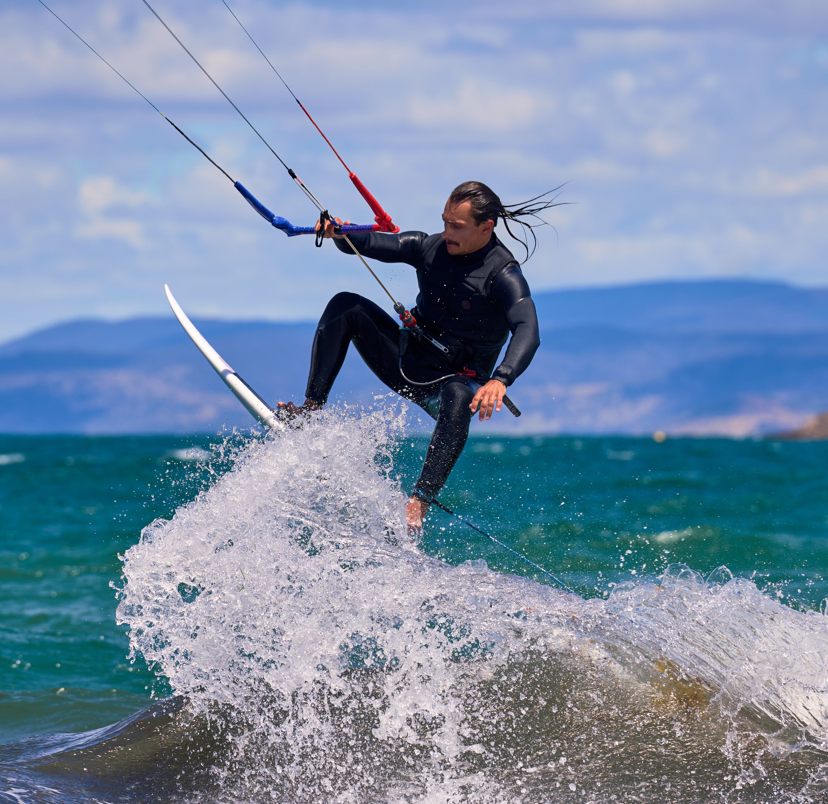 Kitesurfer in black wetsuit riding a crashing wave while holding red kite control lines, with mountains visible across turquoise water.