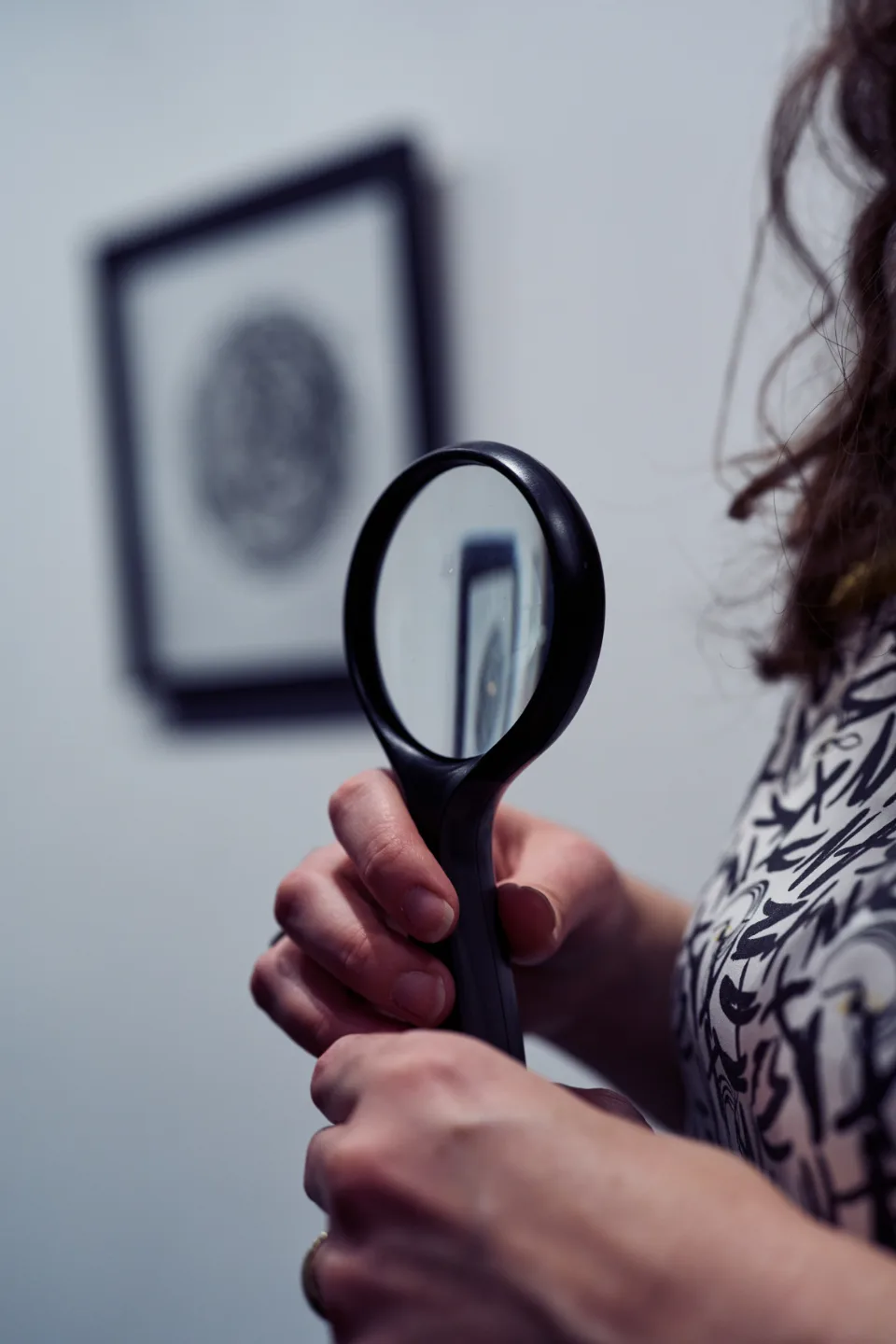 Hand holding magnifying glass with framed portrait visible through the lens and blurred in the background.