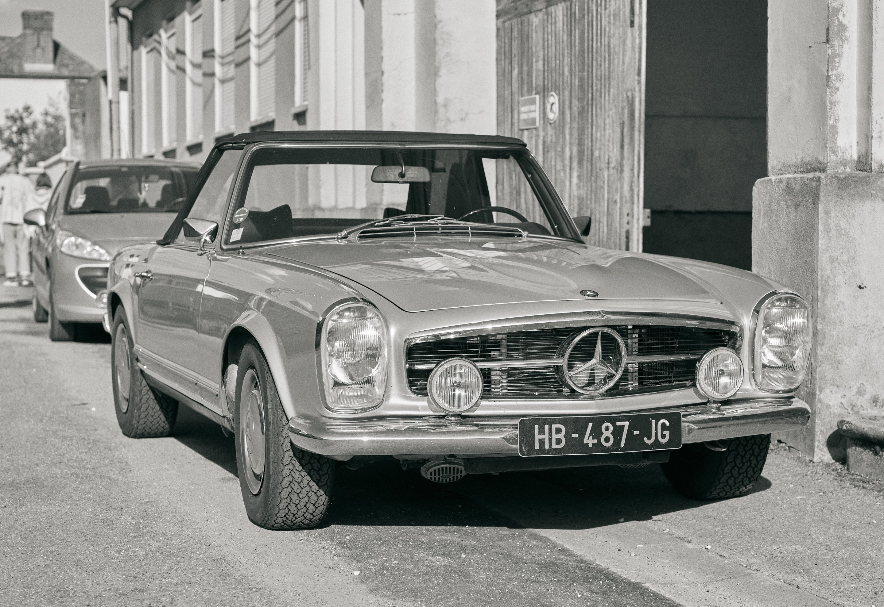 Black and white photograph of vintage Mercedes-Benz convertible parked on street with pillars and faded walls, vintage car visible in background.