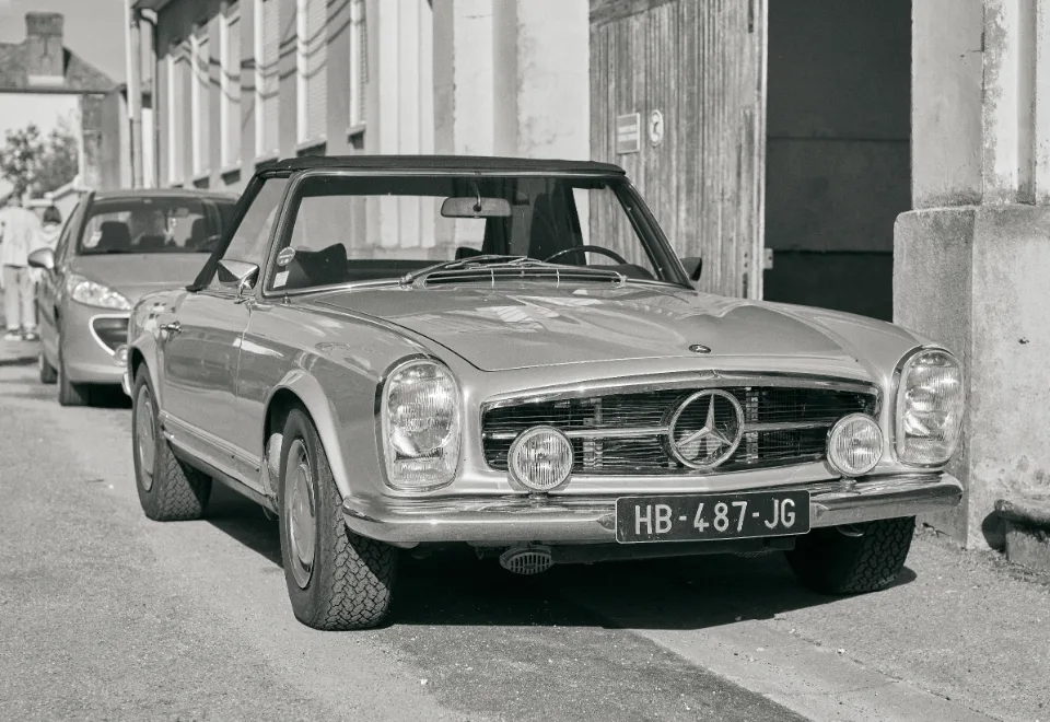 Black and white photograph of vintage Mercedes-Benz convertible parked on street with pillars and faded walls, vintage car visible in background.