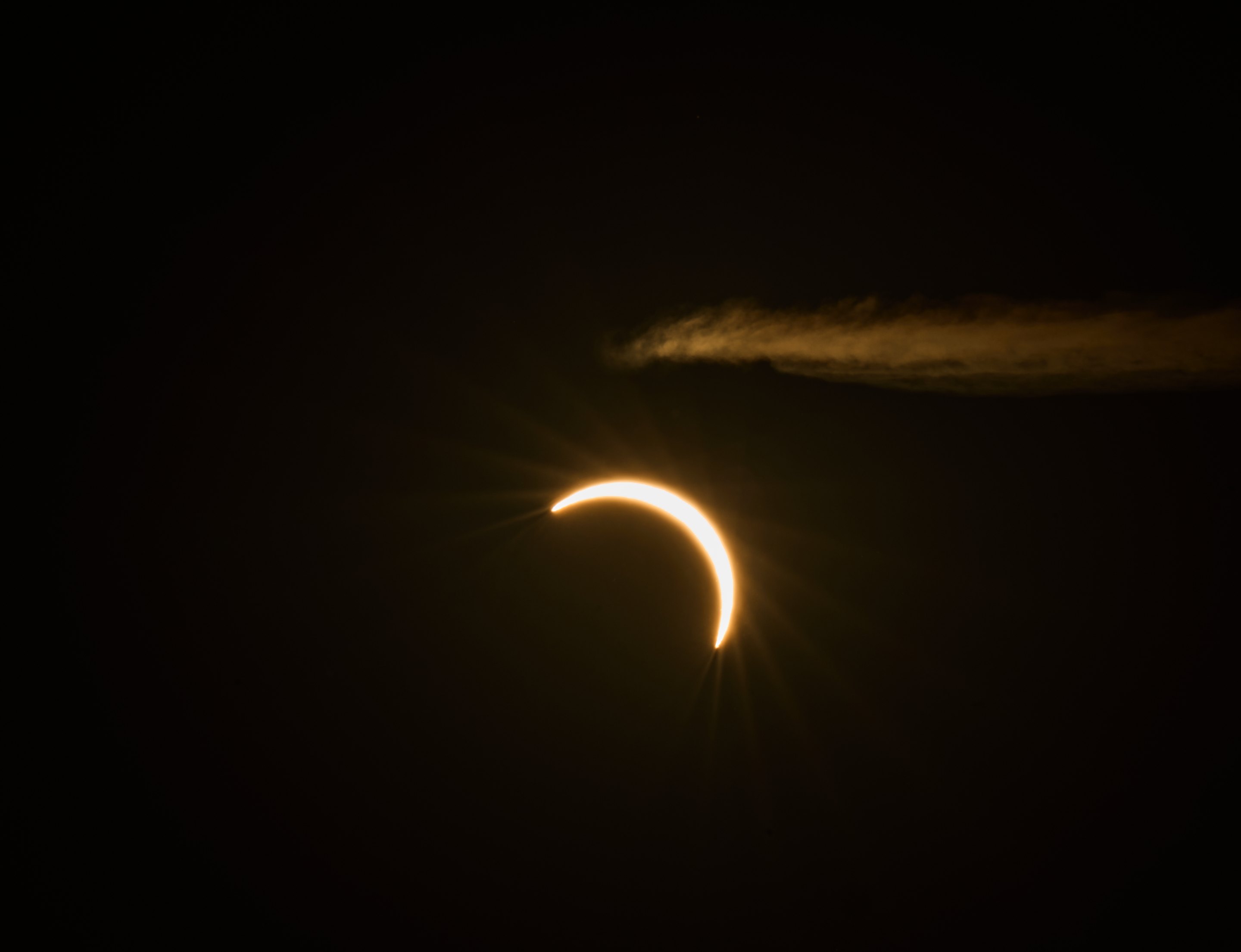 Crescent moon during solar eclipse glowing golden against black sky with aircraft contrail visible above.