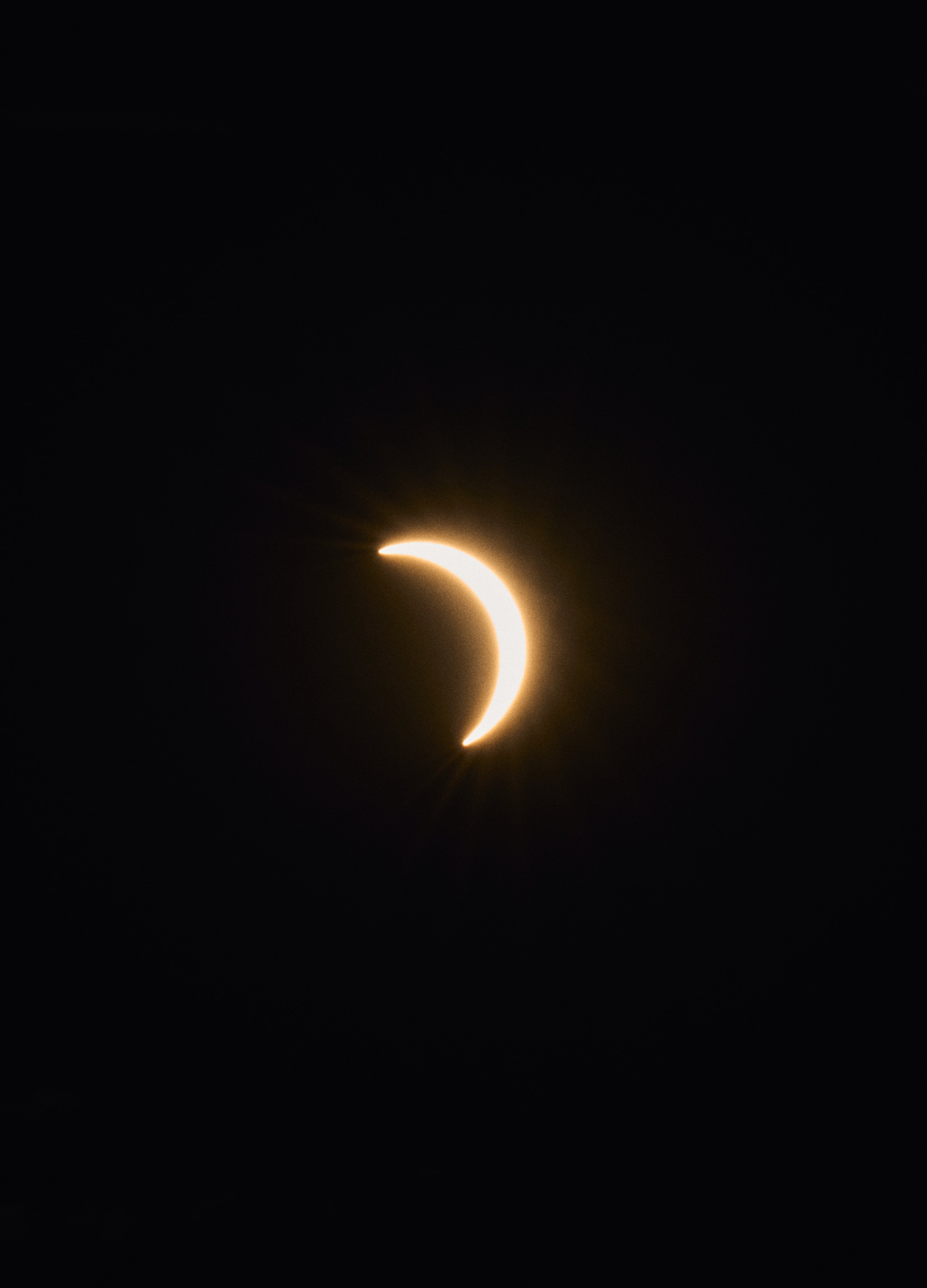 Crescent moon during solar eclipse in vertical frame, bright golden curve against dark sky.